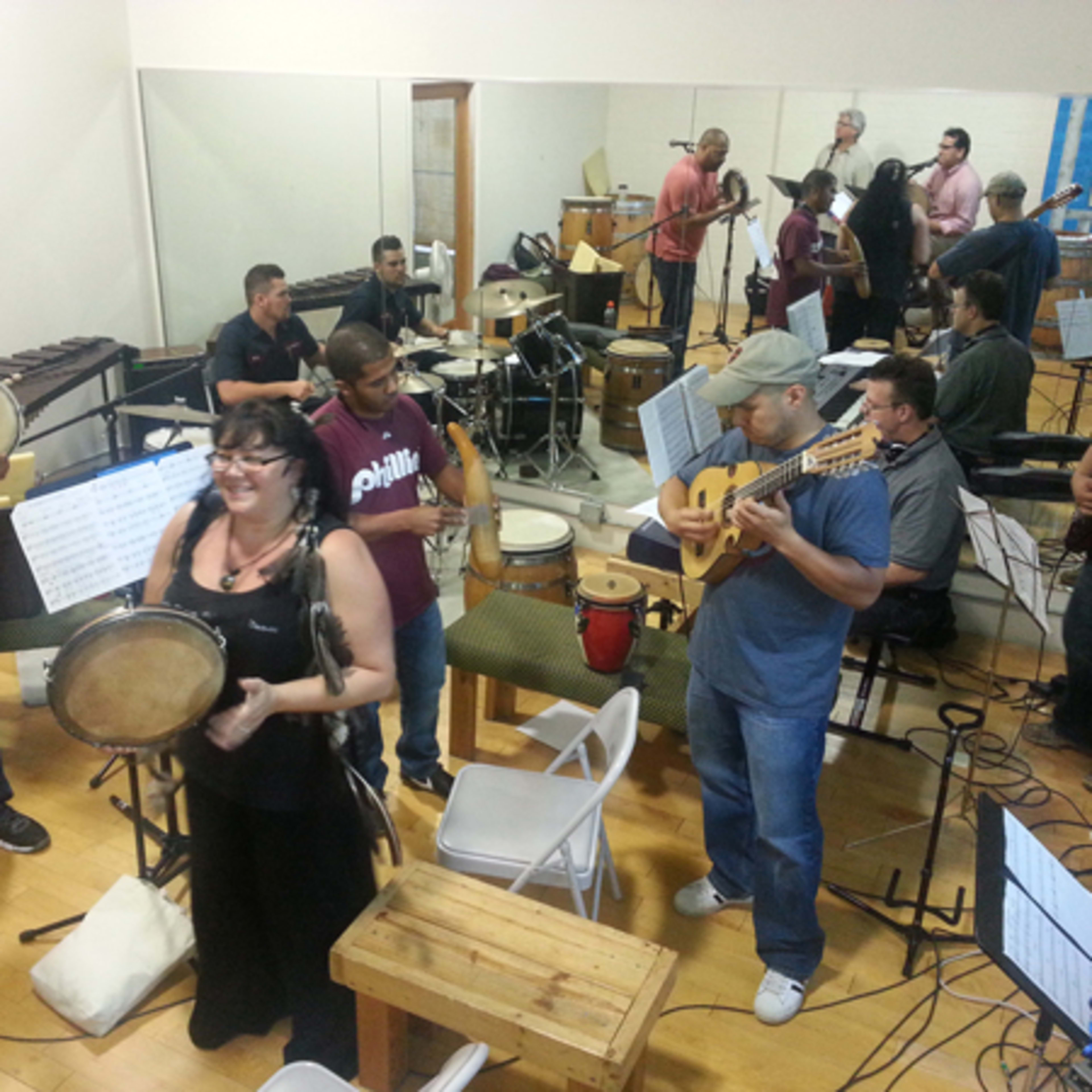 A group of musicians plays various instruments in a rehearsal space filled with chairs and music stands.