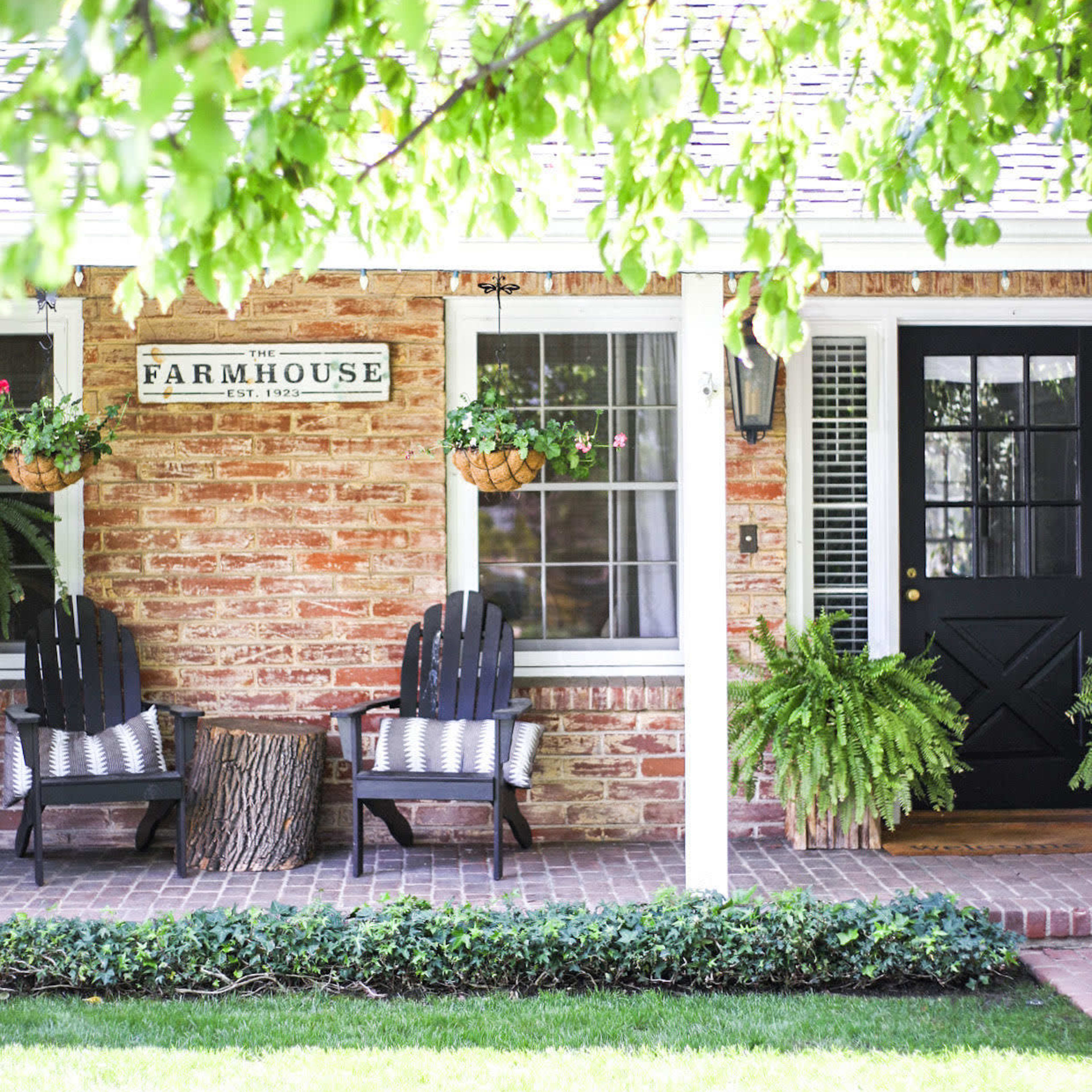 A brick farmhouse with a porch featuring two black chairs, potted plants, and a grassy lawn.