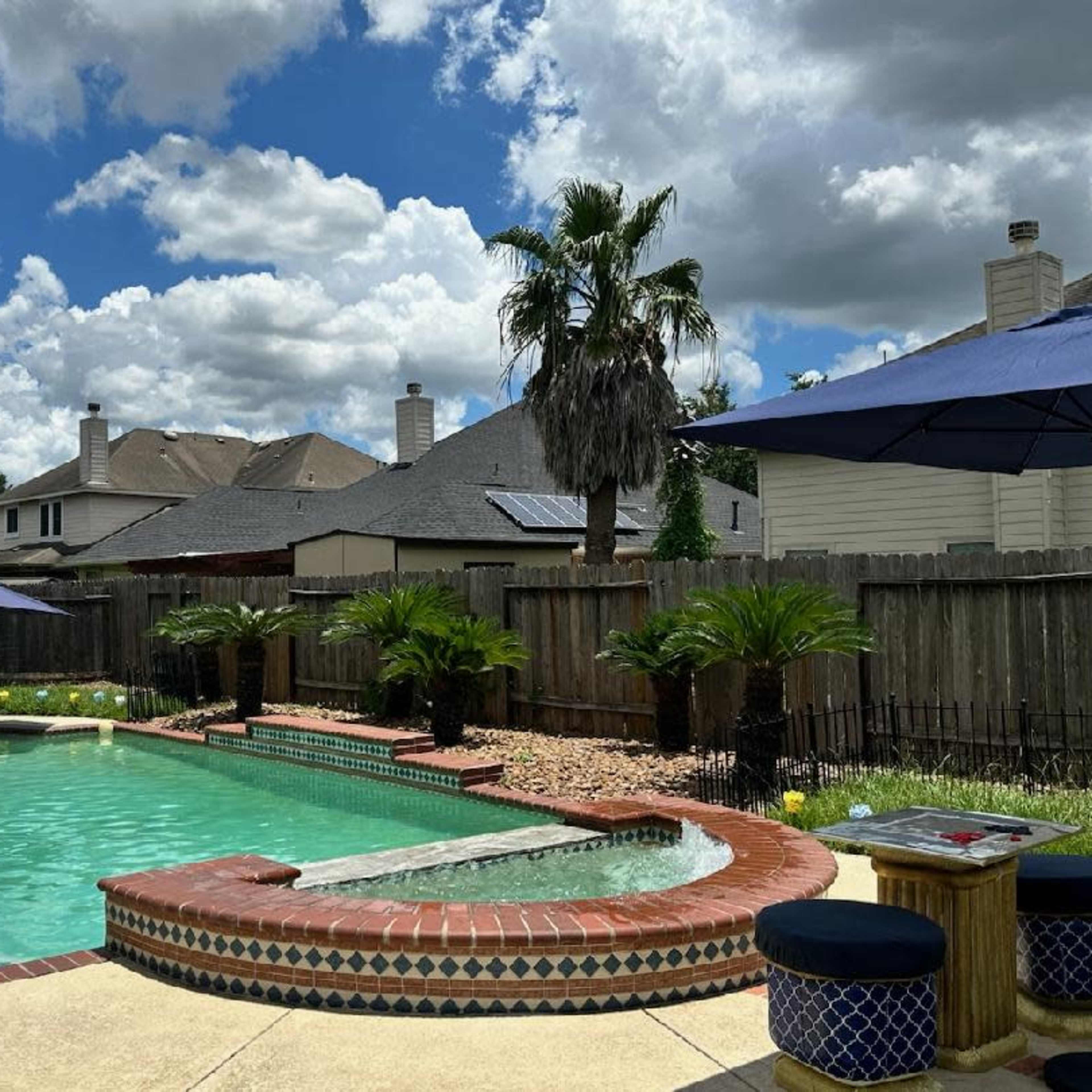 A backyard features a swimming pool with a spa, surrounded by palm trees, umbrellas, and residential homes in the background under a partly cloudy sky.