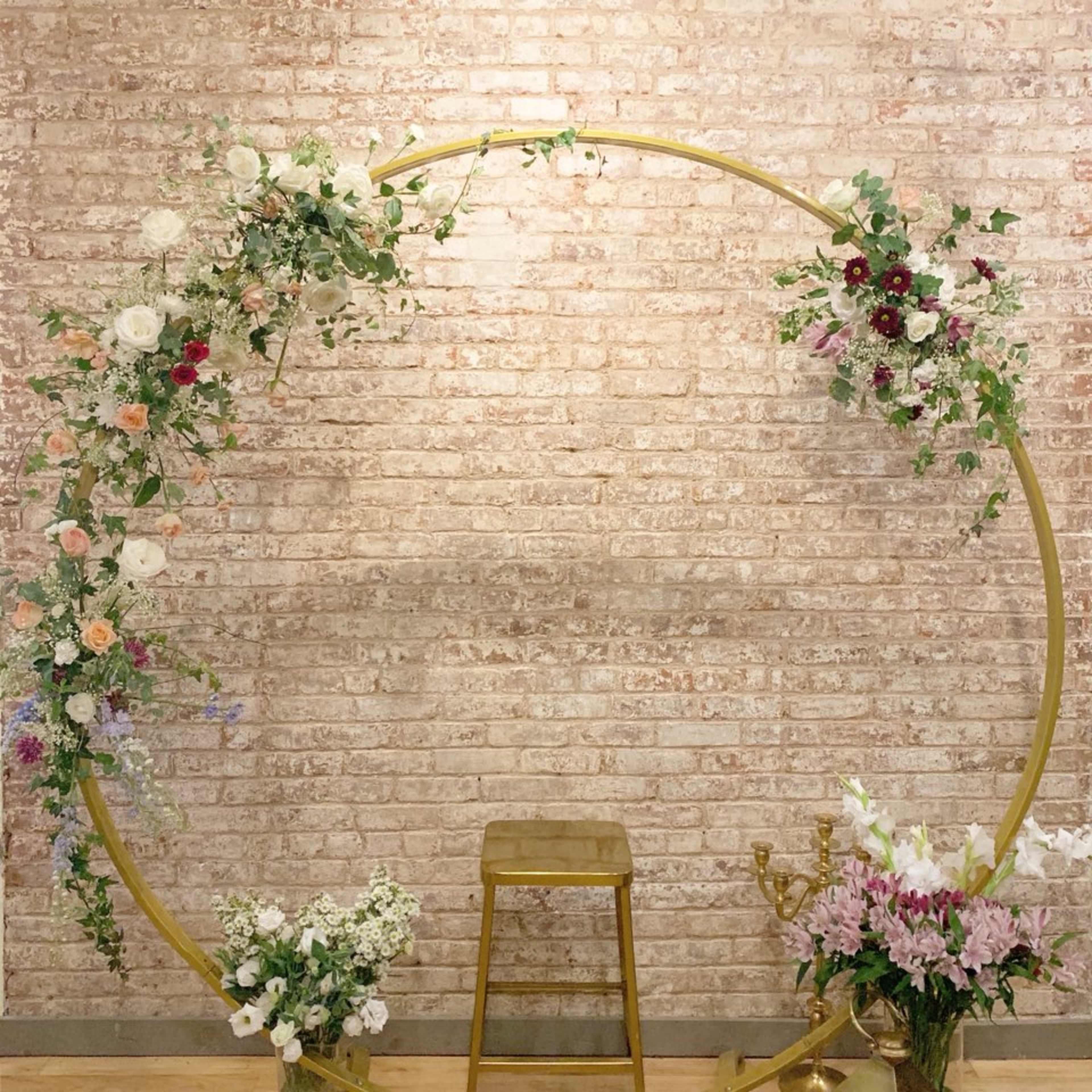 A circular floral arrangement on a brick wall, with a gold stand and a stool positioned in front of it.