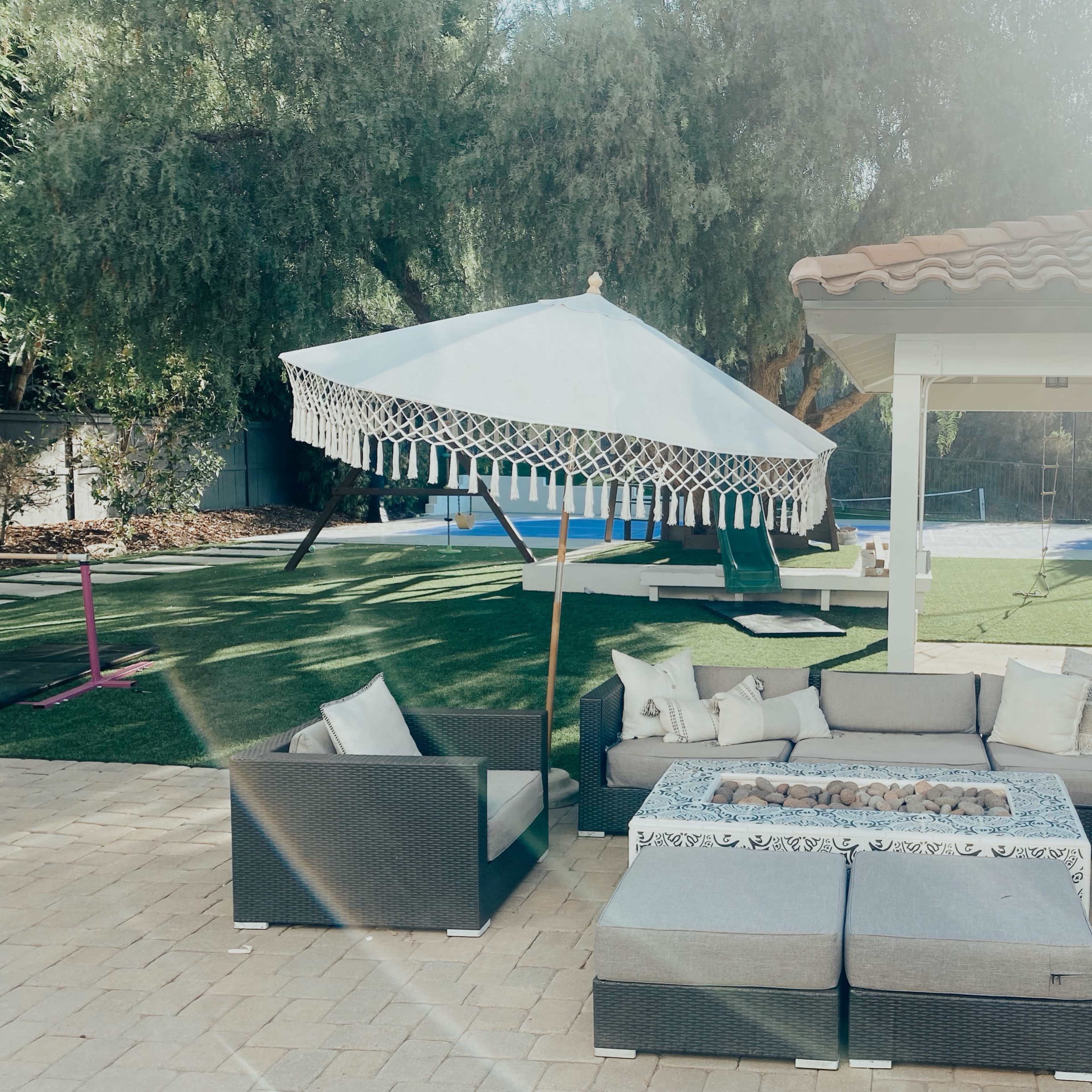 A patio area features a sectional sofa surrounding a fire pit, accompanied by a large umbrella, with a swimming pool and play equipment in the background.