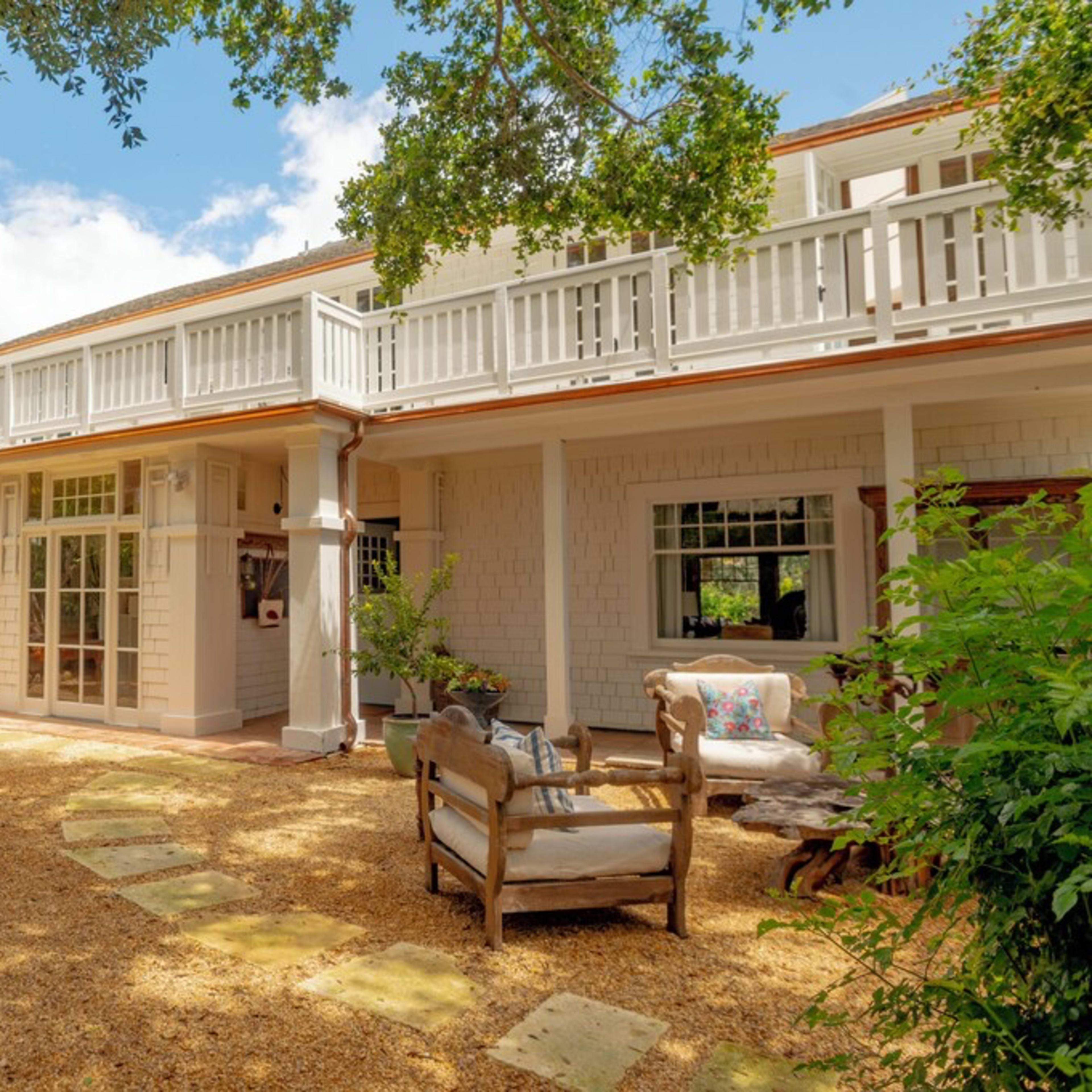A modern two-story house with a sandy yard features a patio and seating area surrounded by greenery.