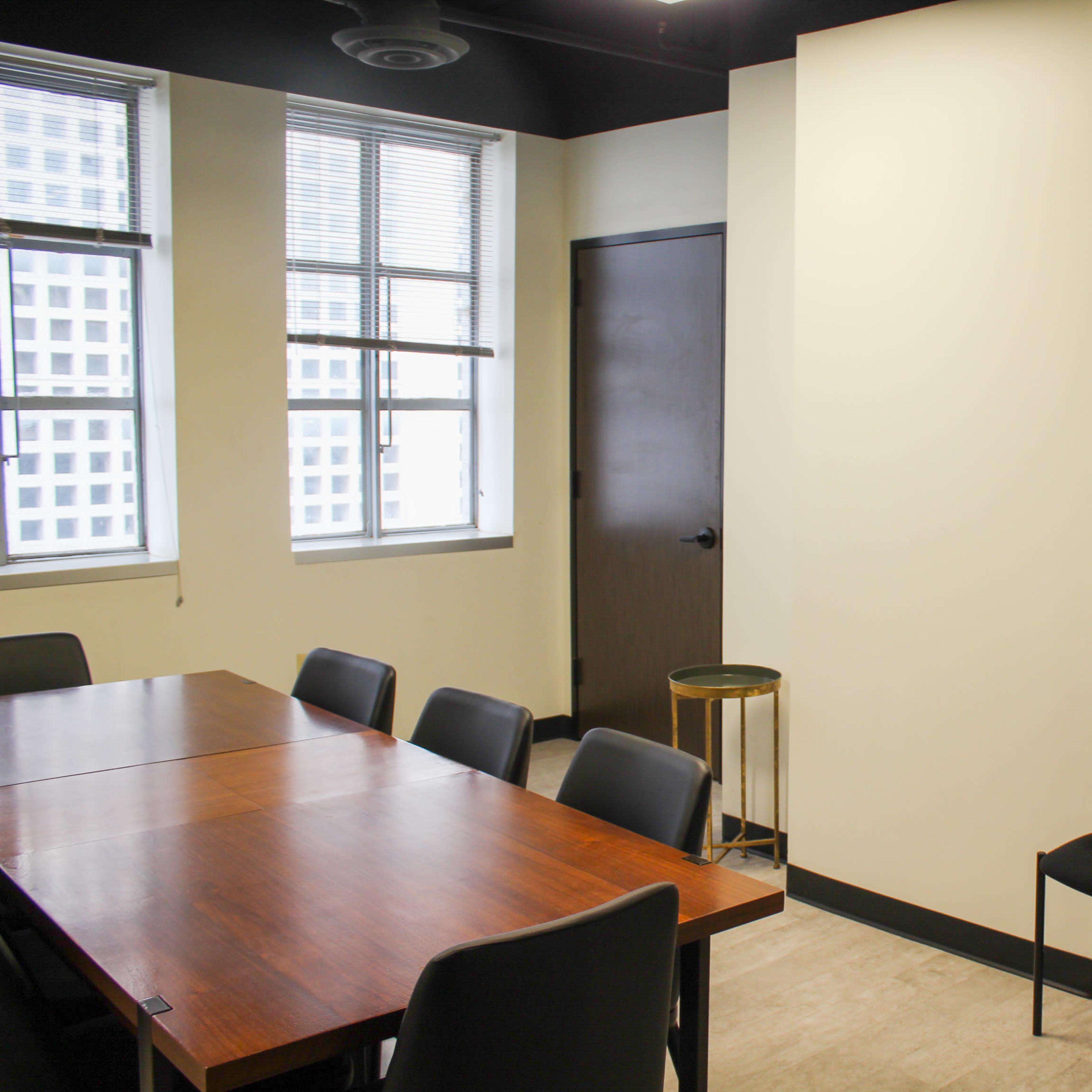 A conference room with a wooden table, several black chairs, a TV mounted on the wall, and large windows allowing natural light.