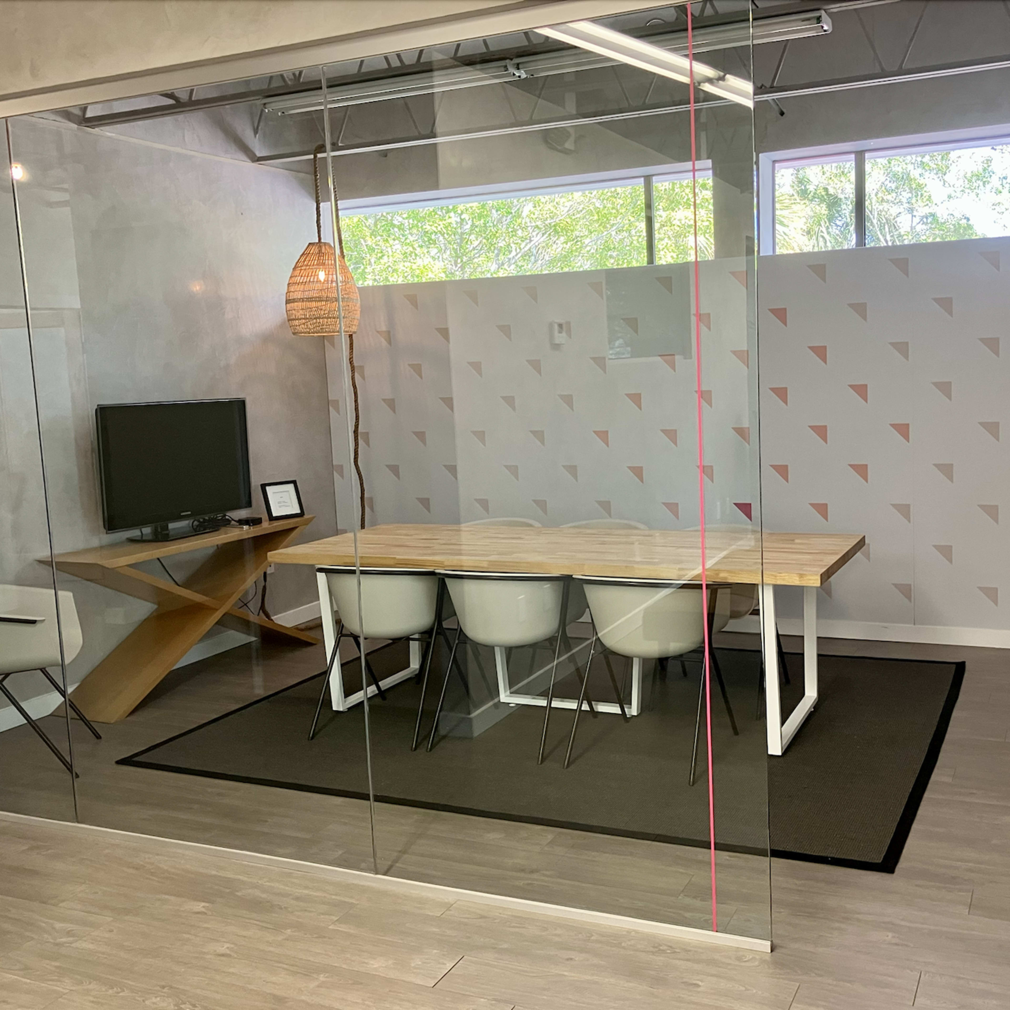 A modern conference room features a wooden table surrounded by white chairs, a television on the wall, and large glass windows with natural light.