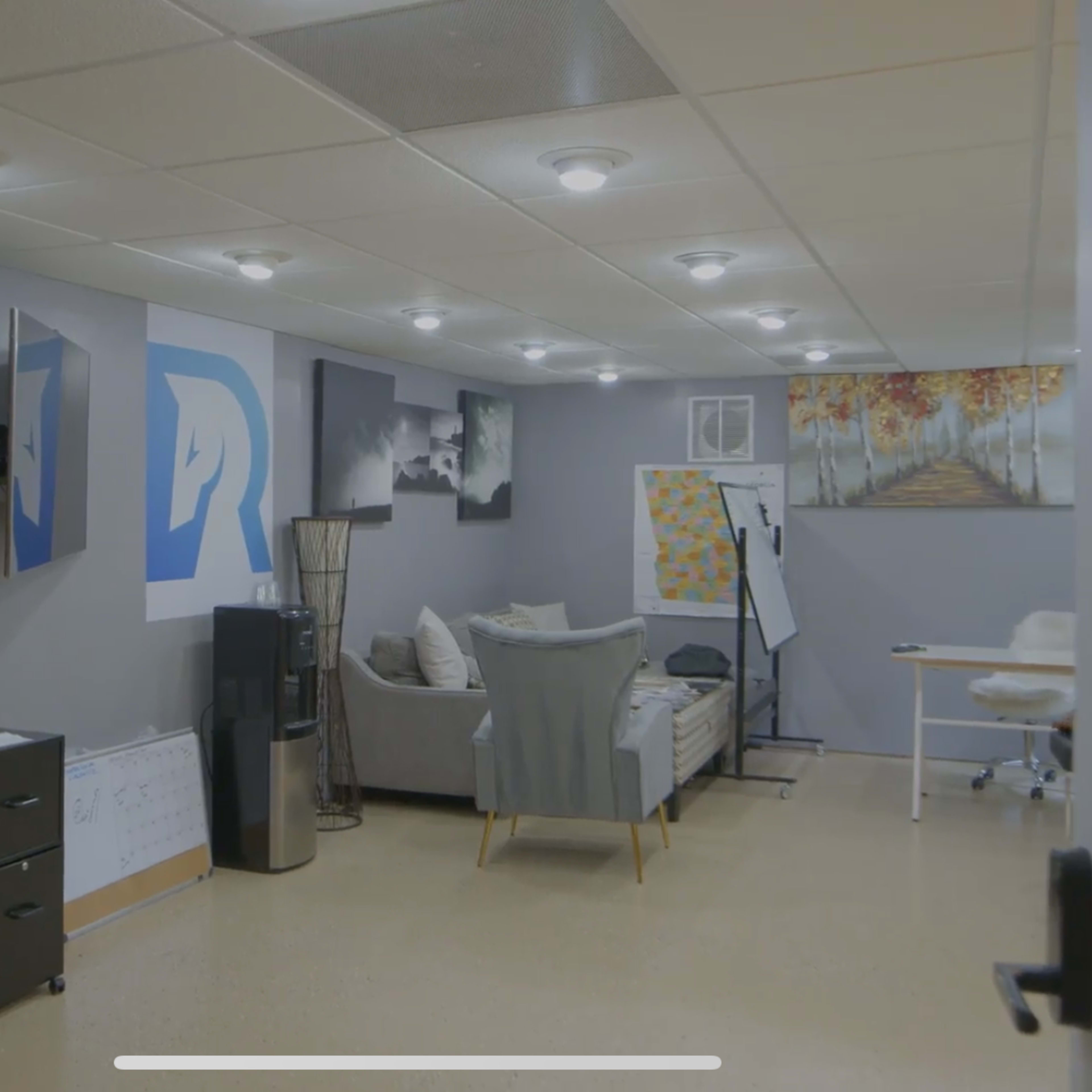A modern office space featuring a gray wall, a sofa, a desk with an ergonomic chair, and various pieces of art on the walls.