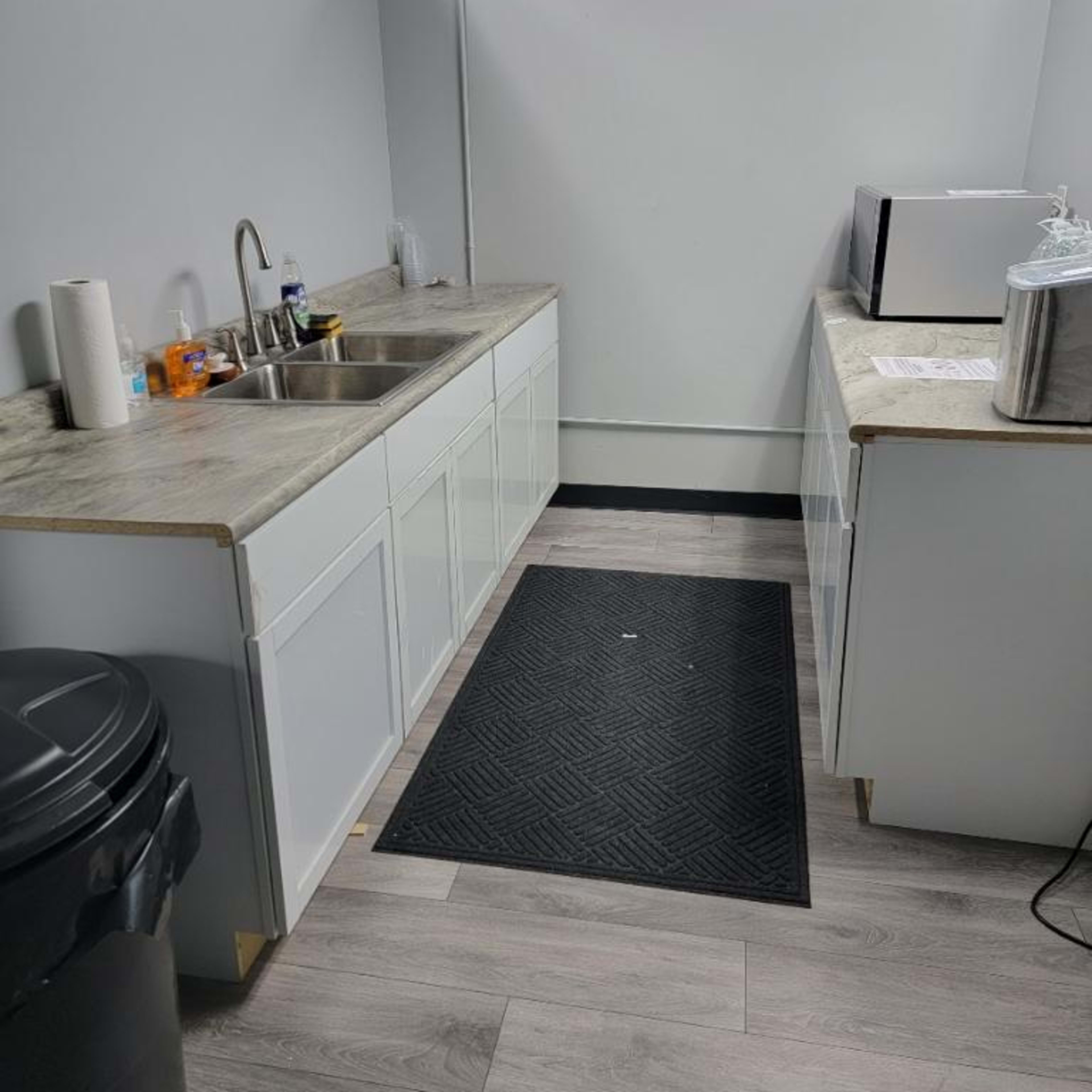 A small kitchen area featuring a sink, countertop, microwave, and a black trash can against a gray wall with light flooring.