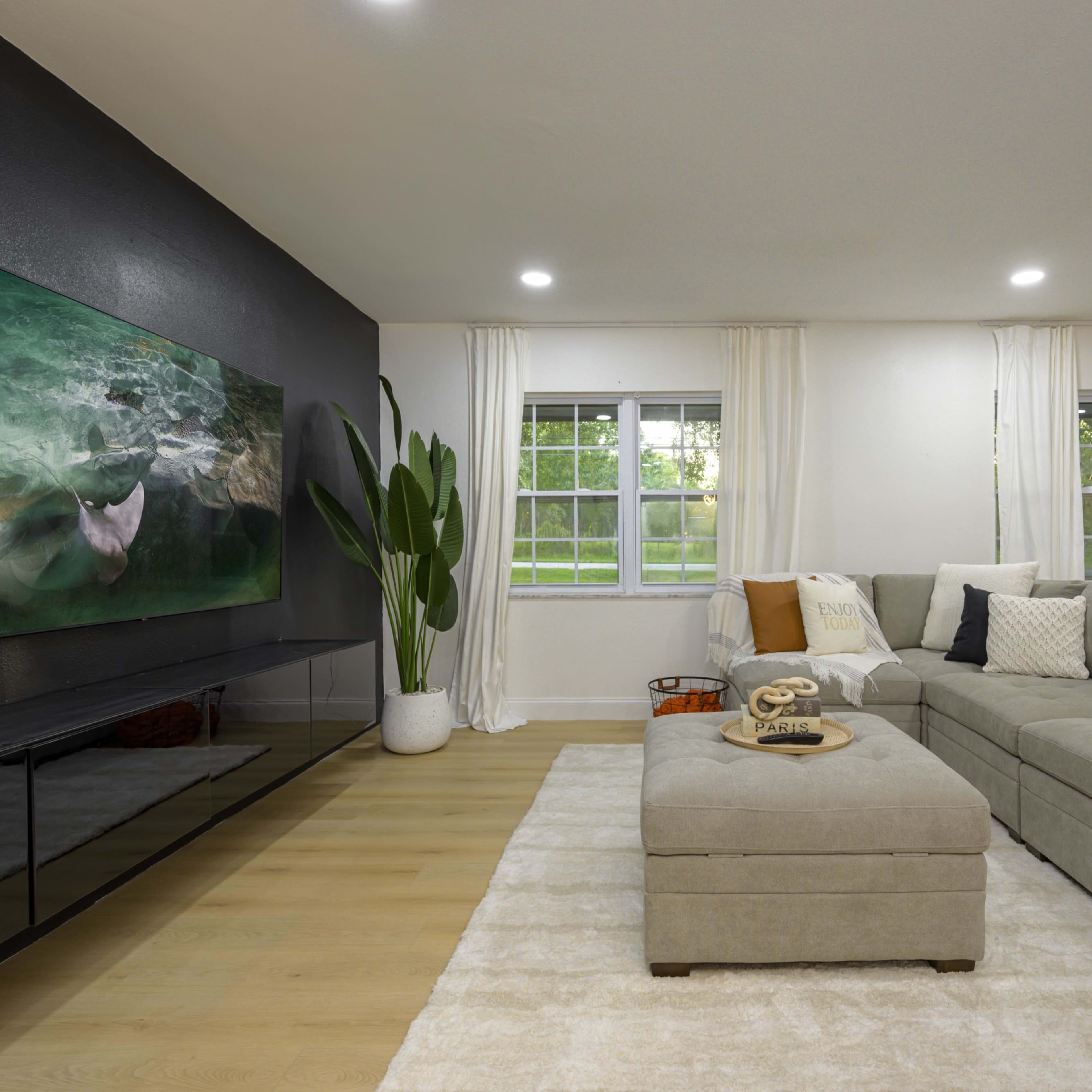 A modern living room with a large sectional sofa, a flat-screen TV mounted on a black accent wall, and large windows allowing natural light.