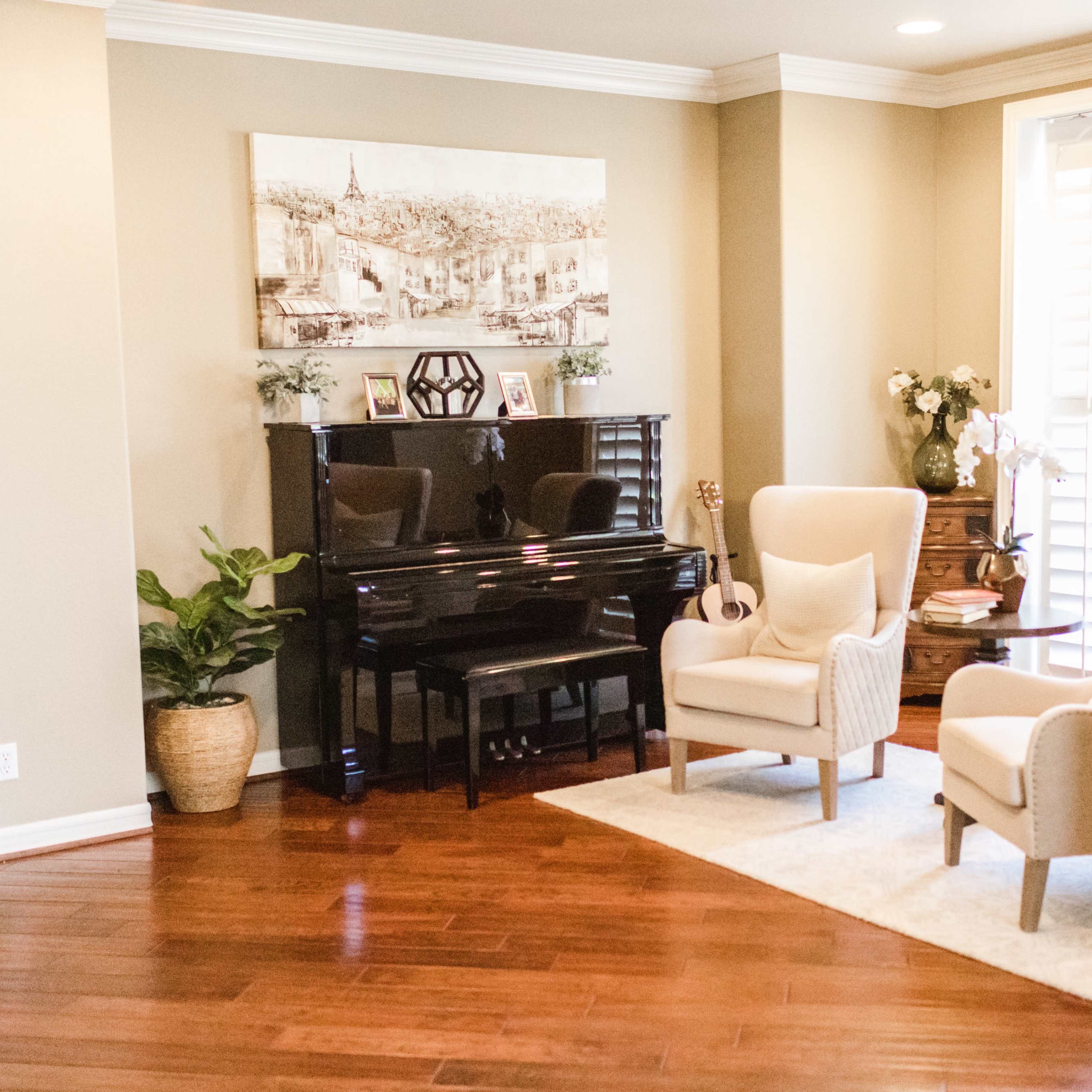The room features a black piano positioned next to two beige armchairs on a light rug, with a decorative plant and a table nearby.