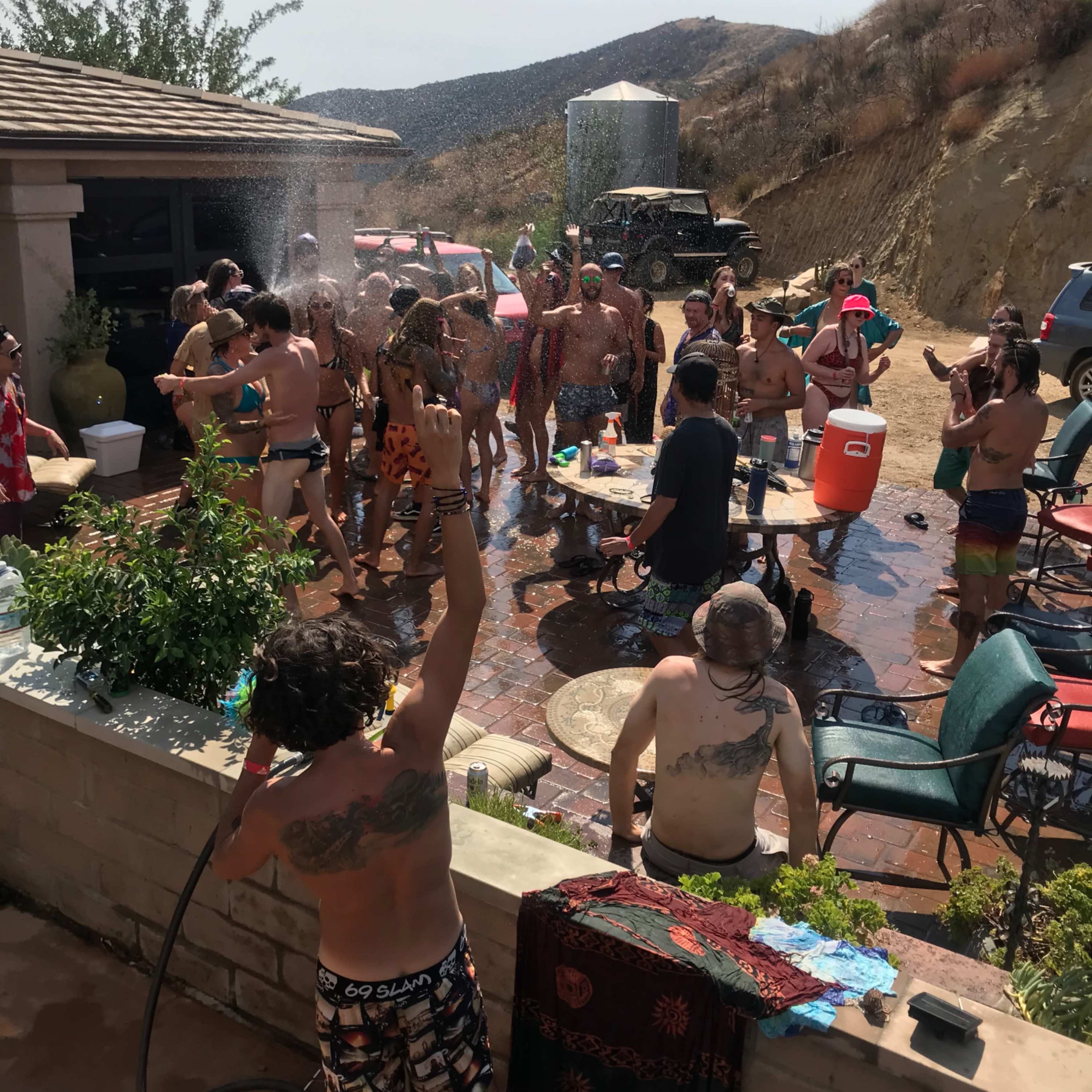 A large group of people are gathered around a poolside area, enjoying a water party on a sunny day with mountains in the background.