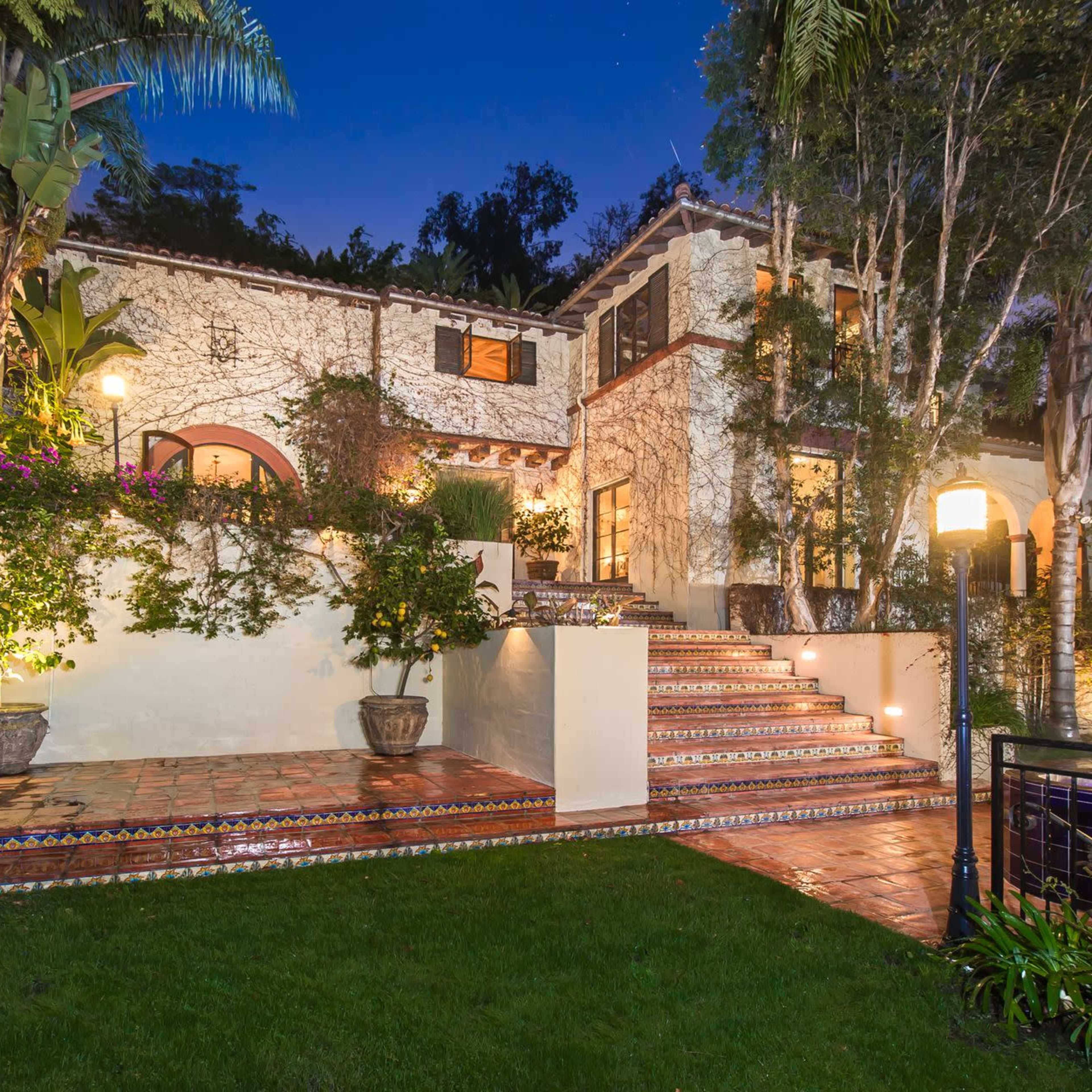 A two-story house with a terraced garden, surrounded by palm trees and illuminated by outdoor lighting.