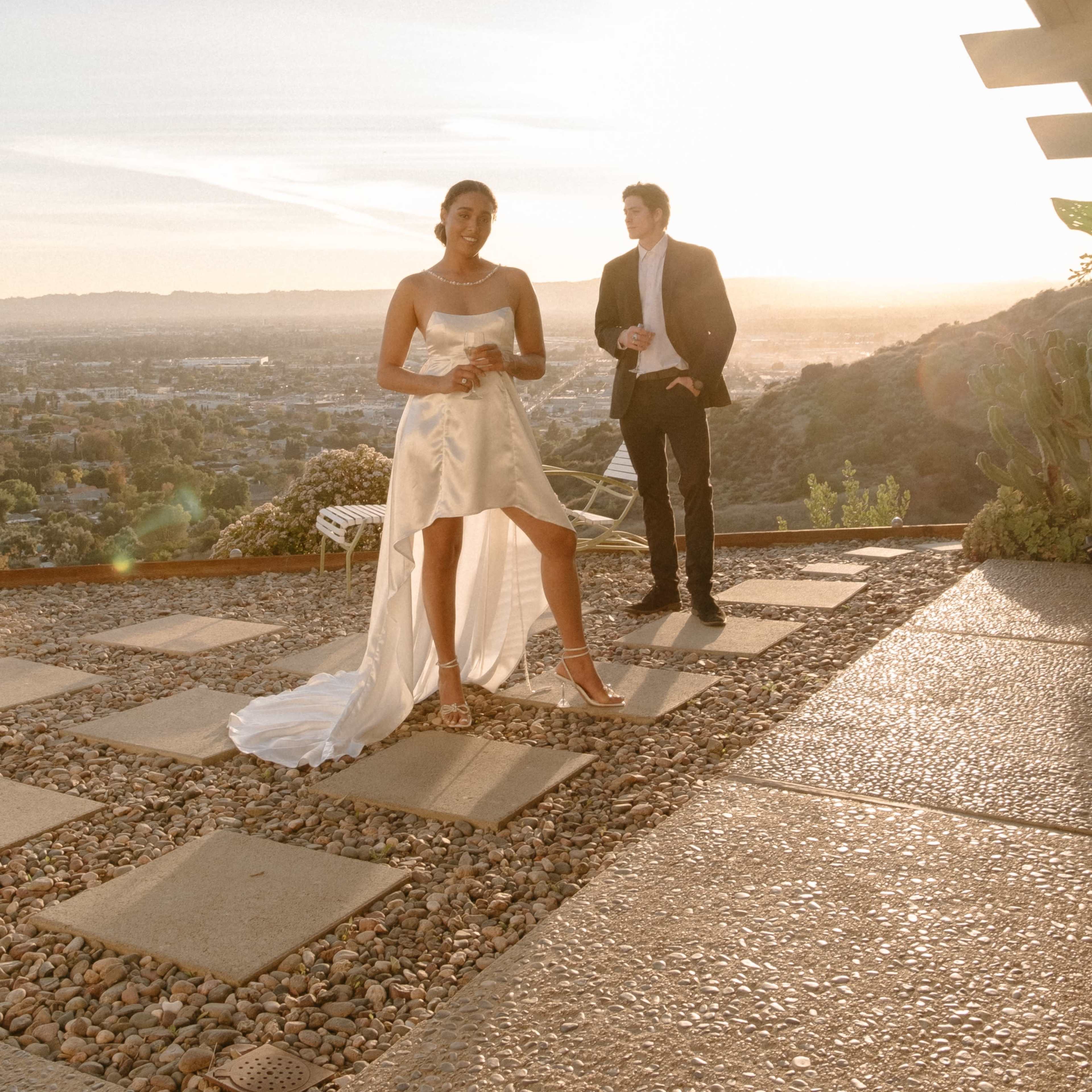 A woman in a strapless white dress stands on a stone patio with a man beside her, overlooking a sunset landscape.
