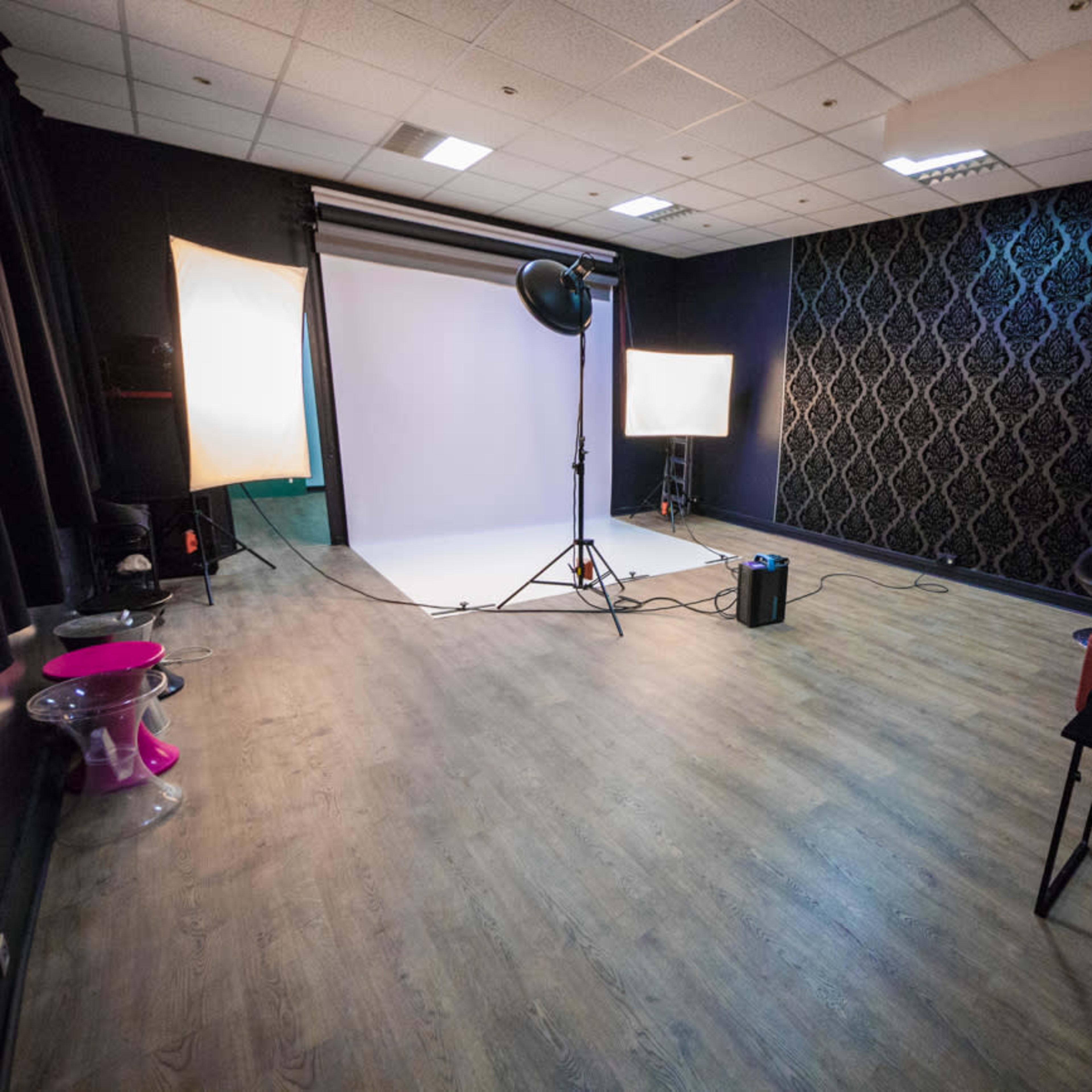 A photo studio with a light setup on one side, a backdrop, and various pieces of furniture scattered around a spacious, wooden floor.