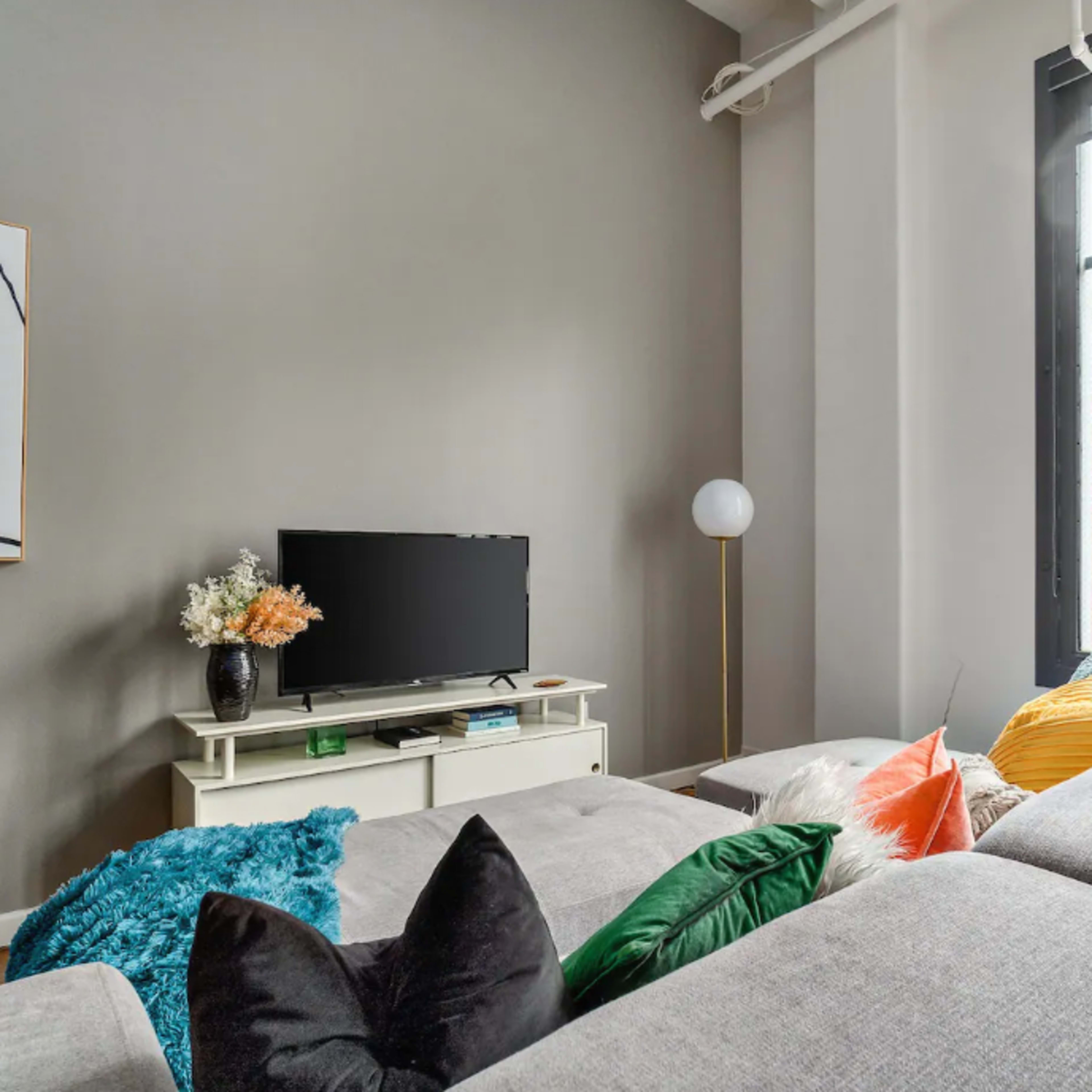 A modern living room with a grey sectional sofa, a television on a light-colored console, and large windows allowing natural light to enter.