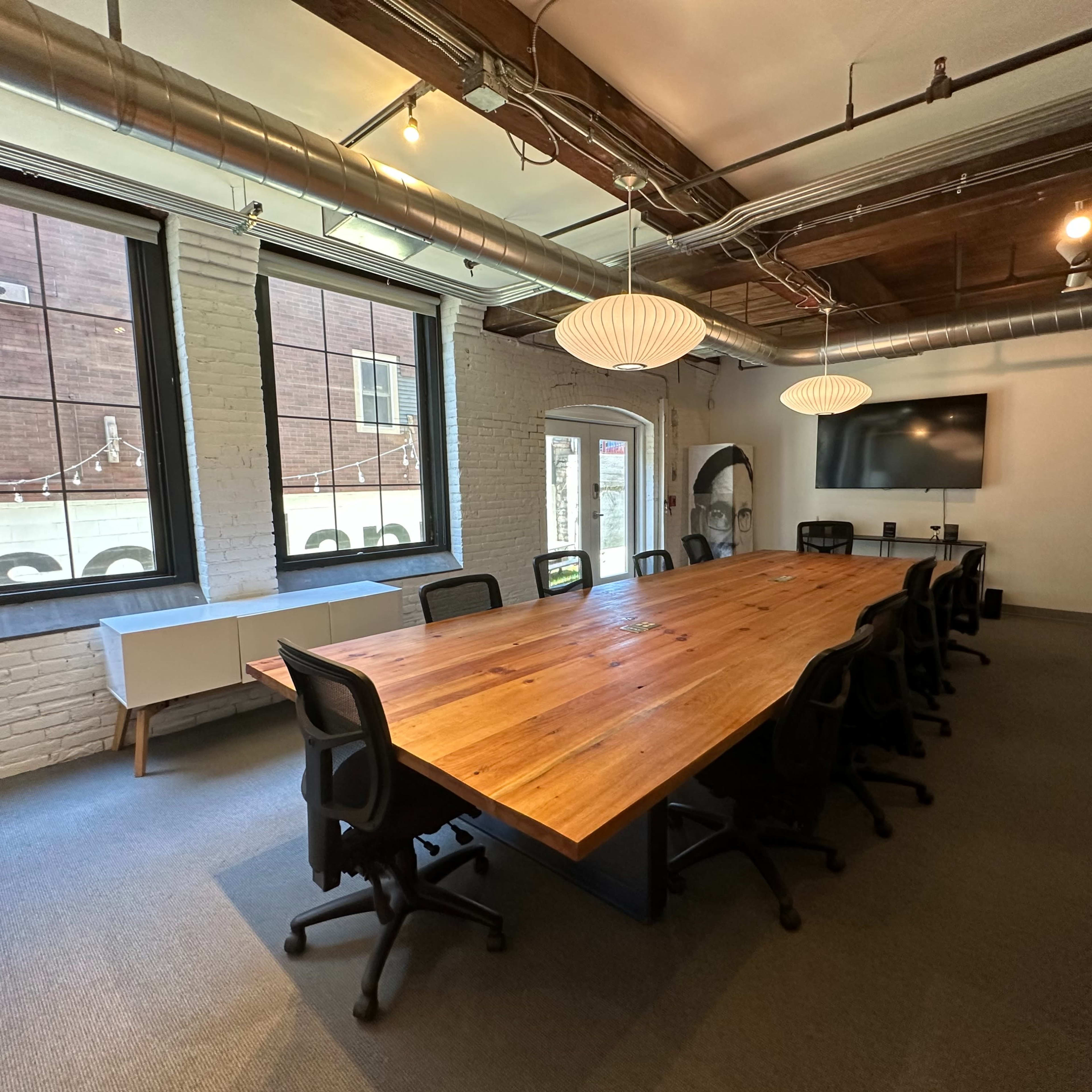 A large wooden conference table with black chairs is centered in a well-lit meeting room featuring exposed brick walls and multiple windows.
