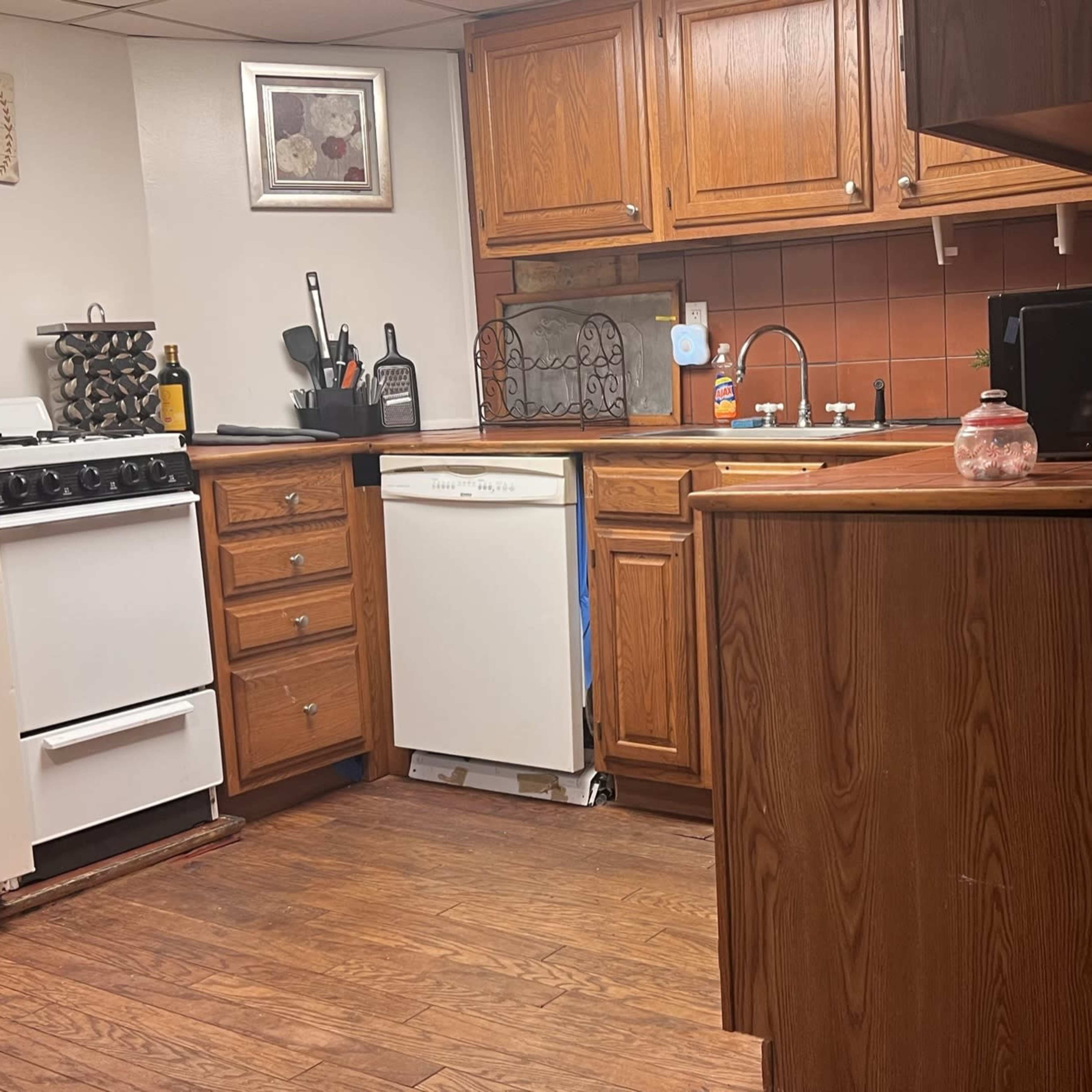 A kitchen with wooden cabinets, a white refrigerator, and a dishwasher, featuring a brown tiled backsplash and hardwood flooring.