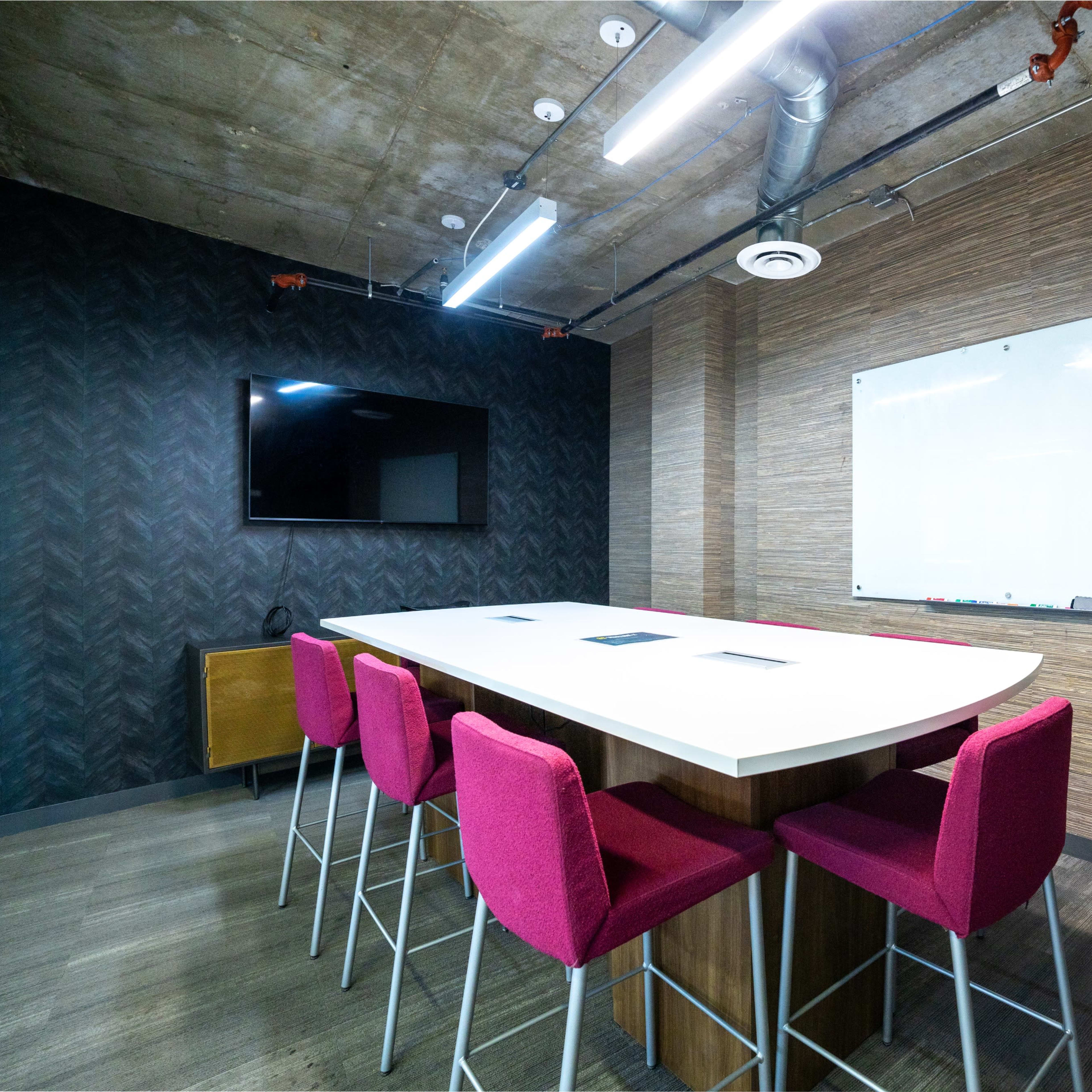 A modern meeting room features a long, white table surrounded by pink bar stools, a flat-screen TV, and a whiteboard on a textured wall.