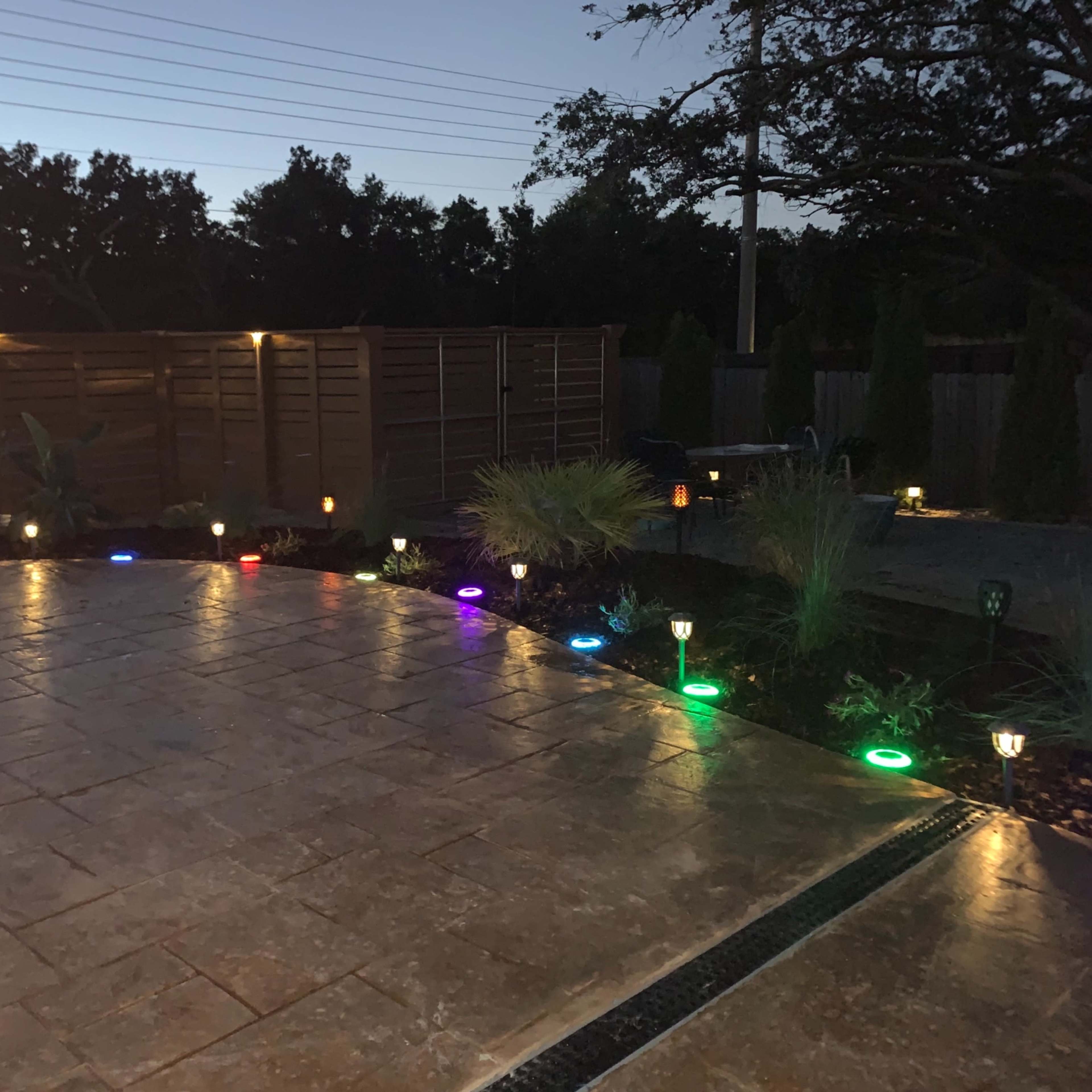 A patio area illuminated by colorful lights along the edges, surrounded by plants and a wooden fence.