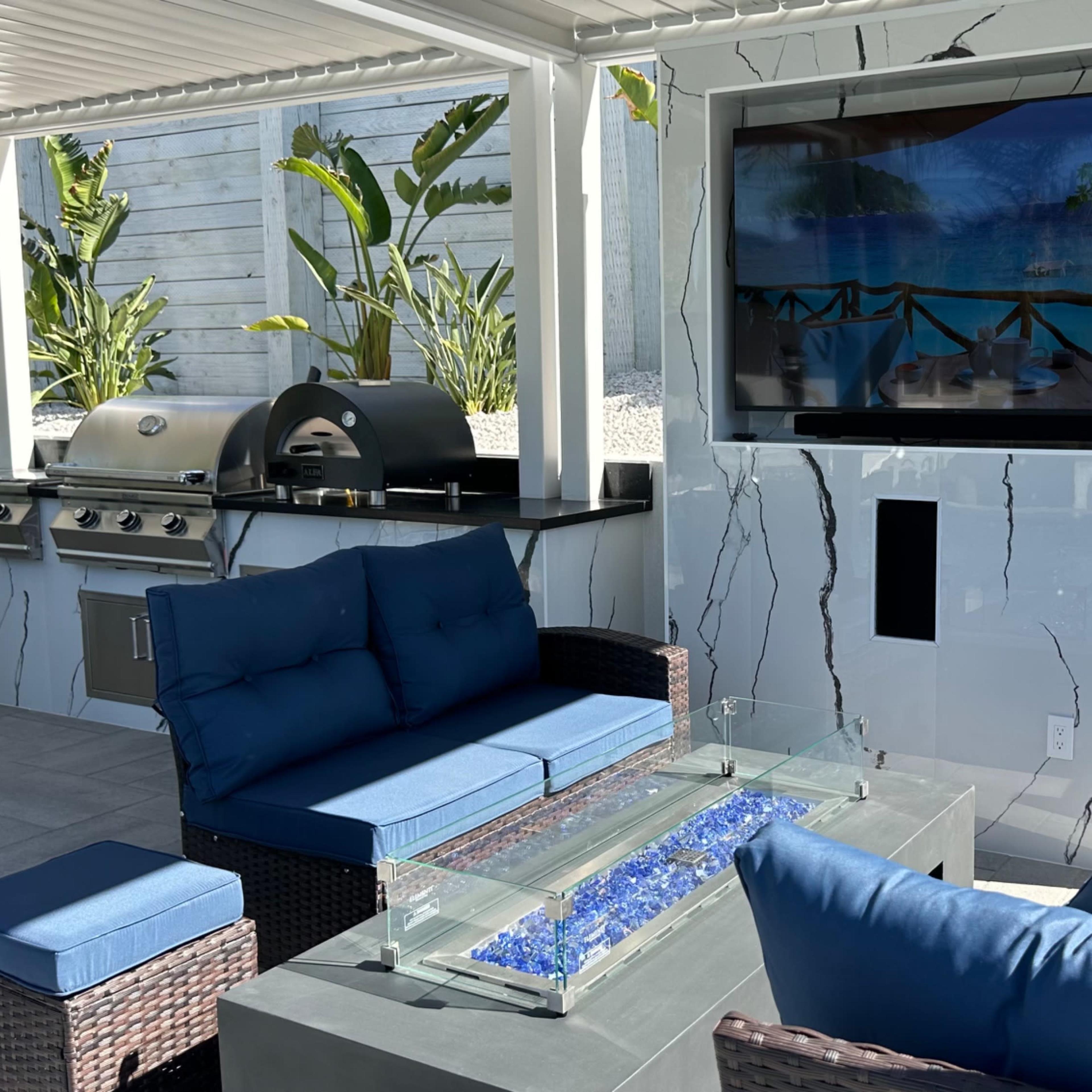 A modern outdoor living space features a sleek kitchen with a grill, a flat-screen TV mounted on a marble wall, and blue cushioned seating with a glass-top coffee table.