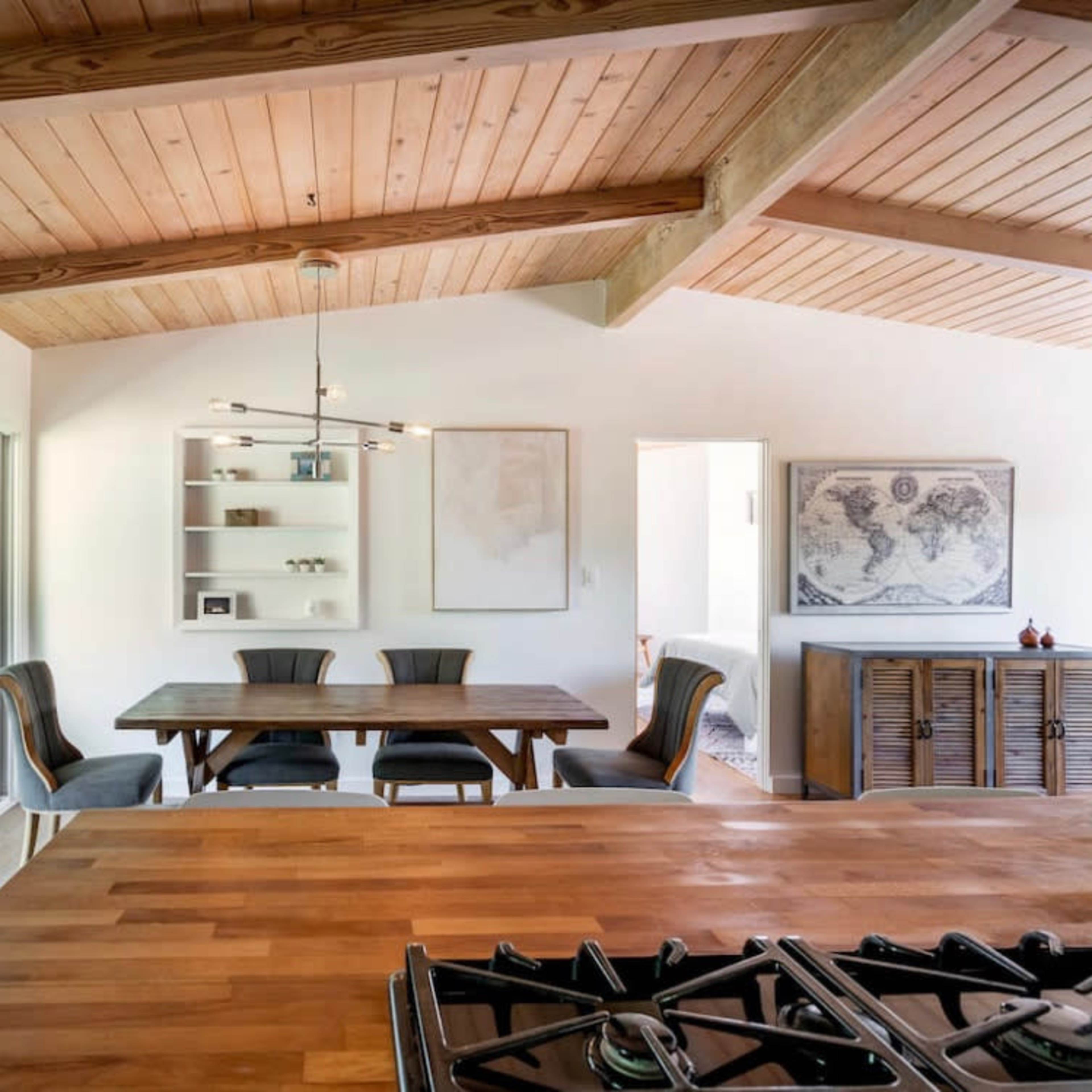 A modern dining area features a wooden table and chairs beneath a slanted wooden ceiling, with large windows that overlook an outdoor space.