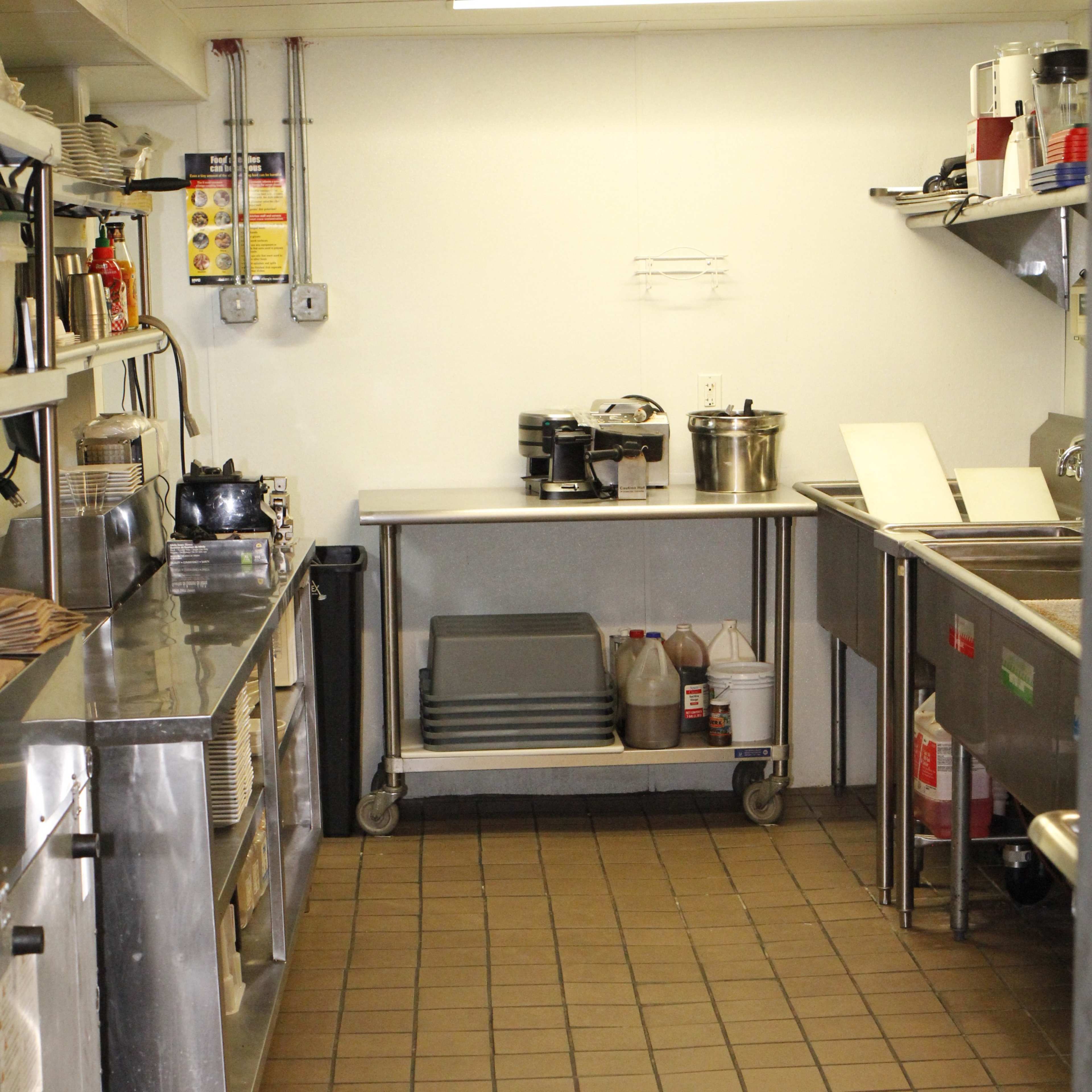 A commercial kitchen with stainless steel countertops, shelves, and various kitchen equipment arranged along the walls and a tiled floor.