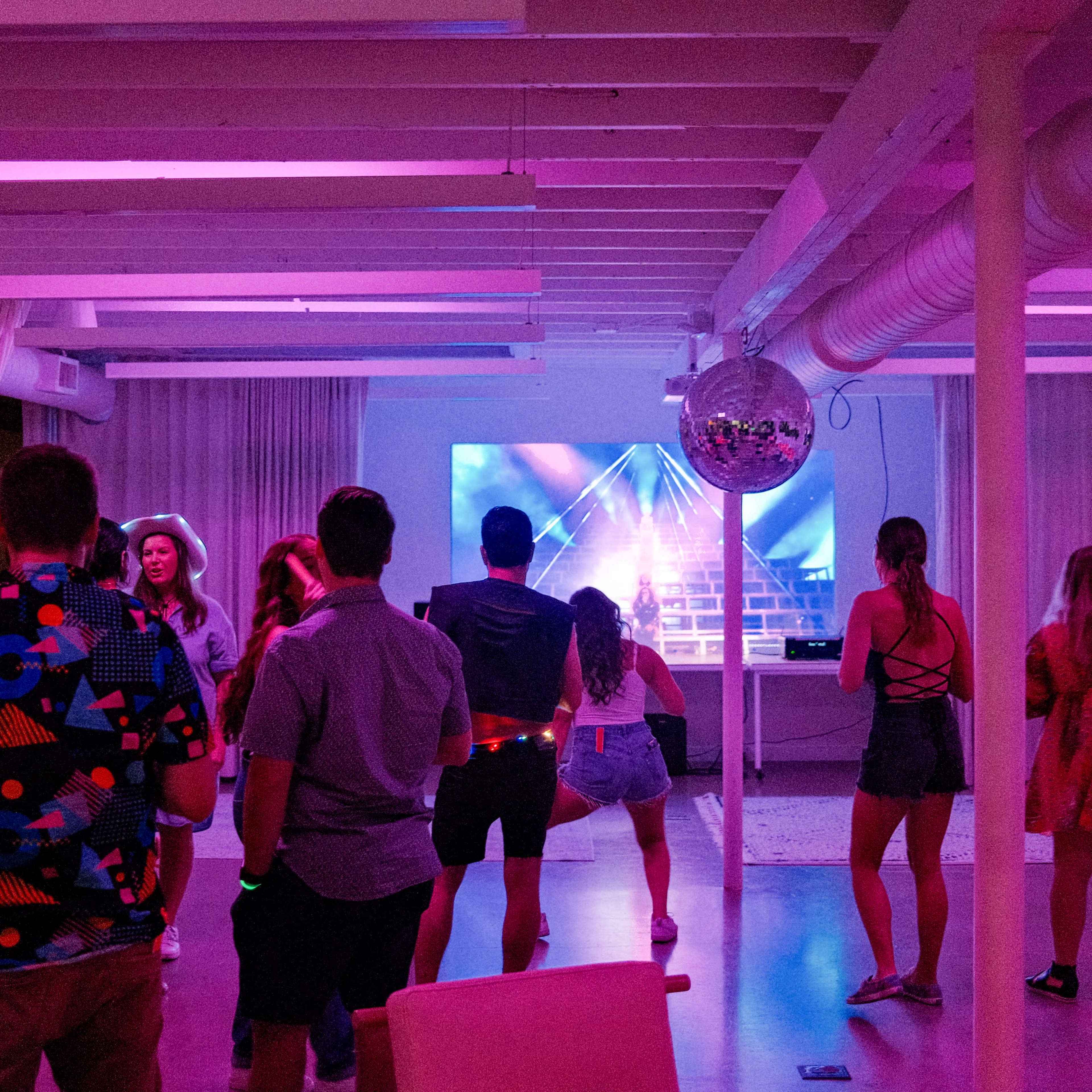 A group of people dance in a brightly lit room with colorful lighting and a large screen displaying a performance.