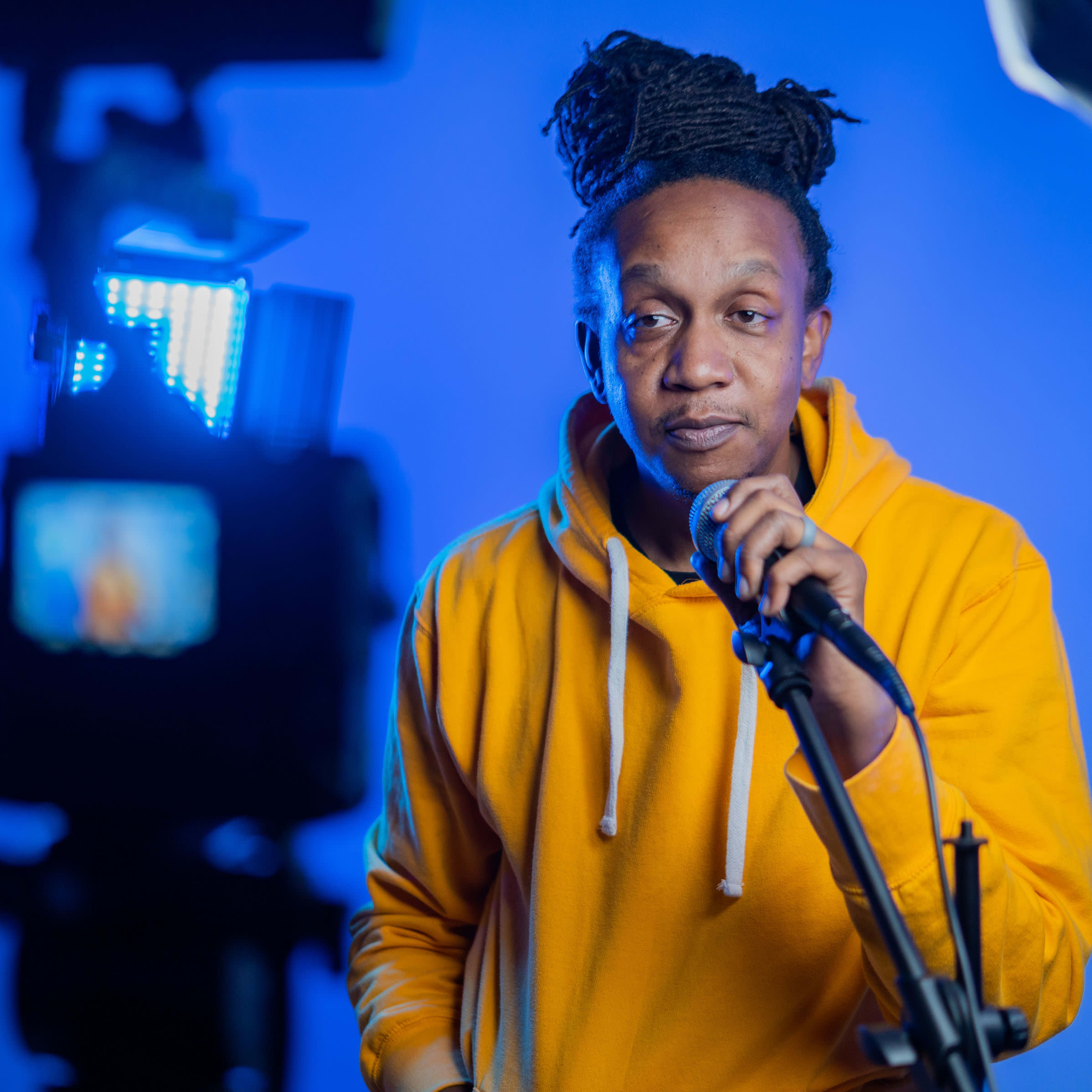 A person in a yellow hoodie stands in front of a microphone, speaking into it while being illuminated by a blue backdrop and studio lights.