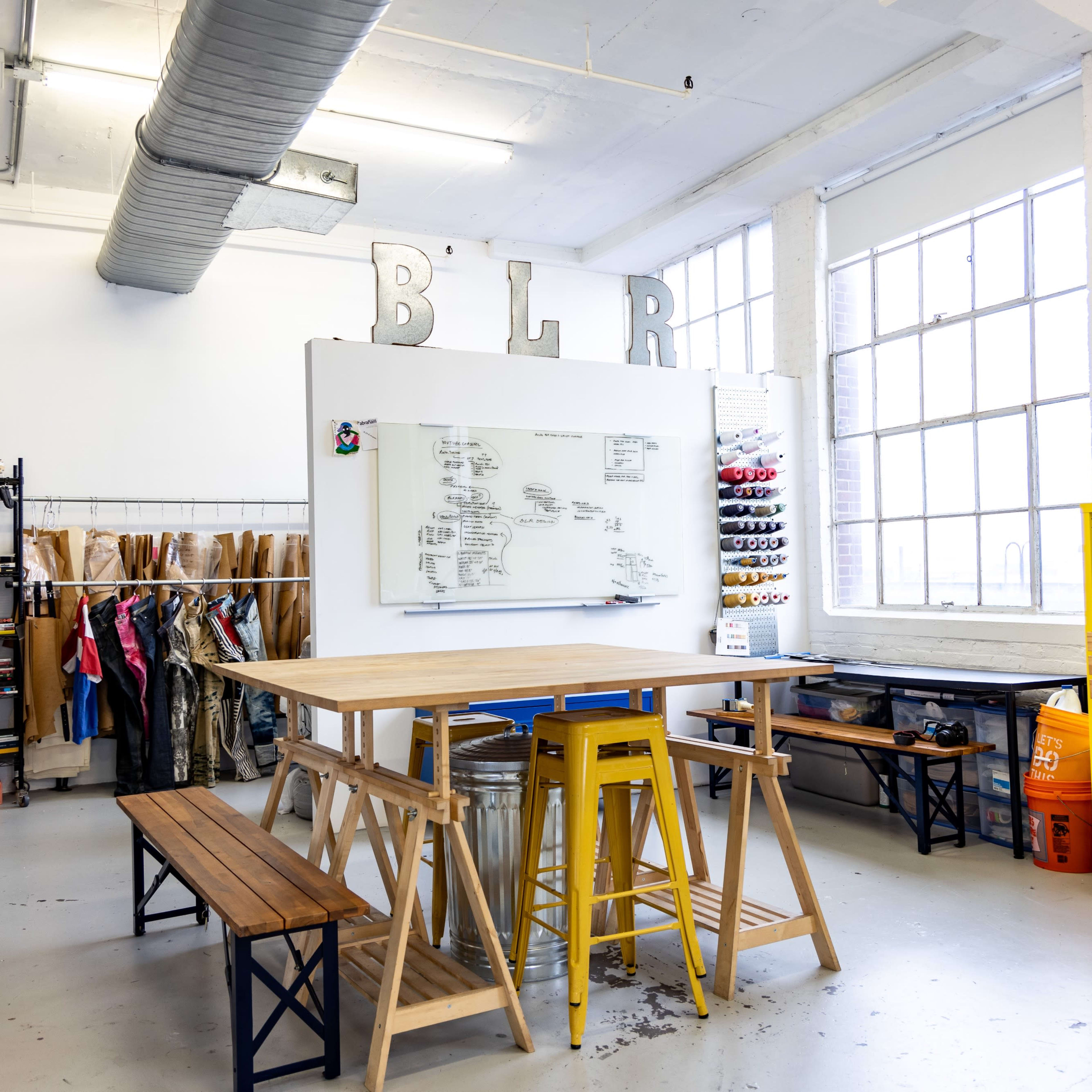 A creative workspace features a large table, colorful storage boxes, and a whiteboard, with shelves filled with books and materials in a well-lit room.