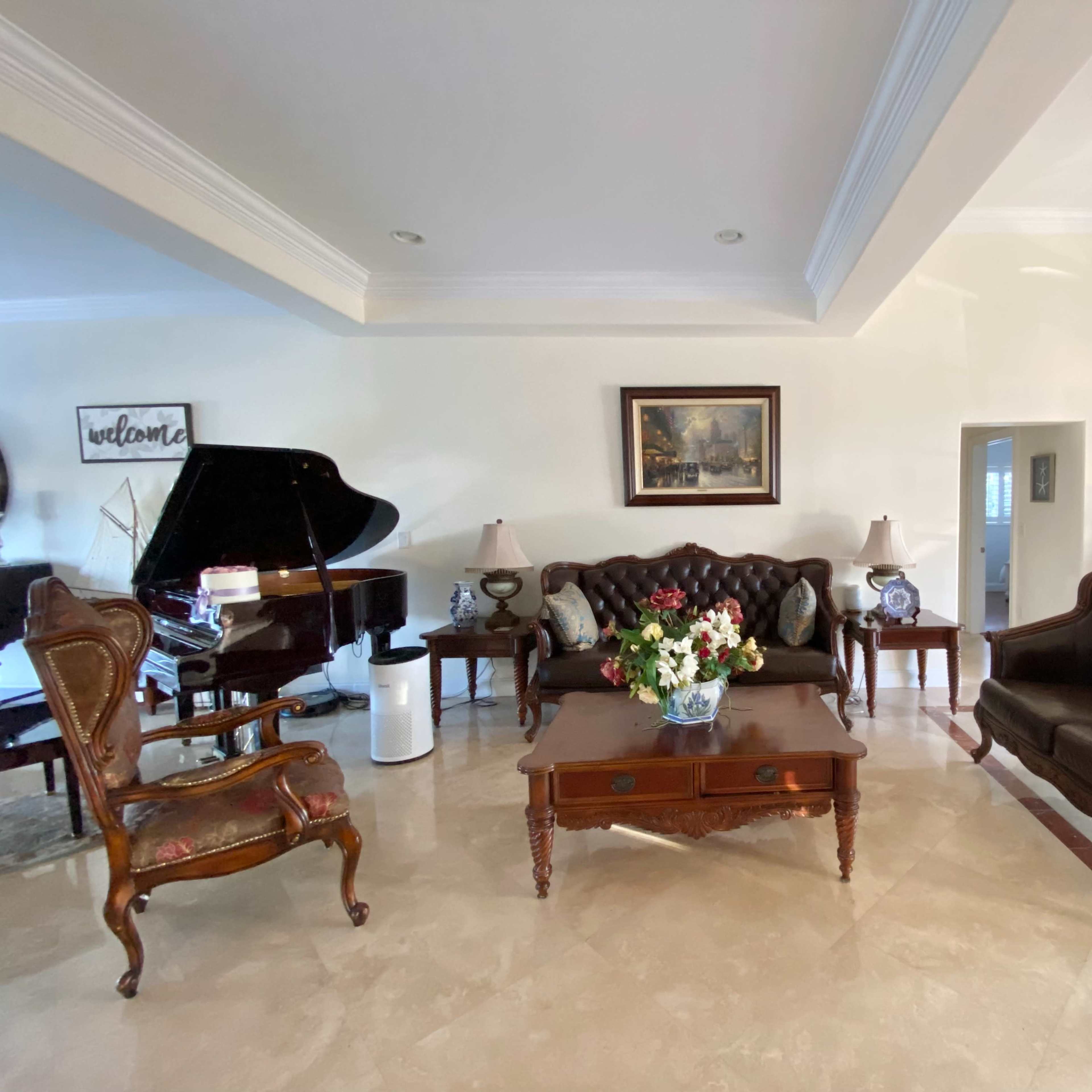 The room features a grand piano, two upholstered sofas, a wooden coffee table, and decorative lamps, arranged around a floral centerpiece.