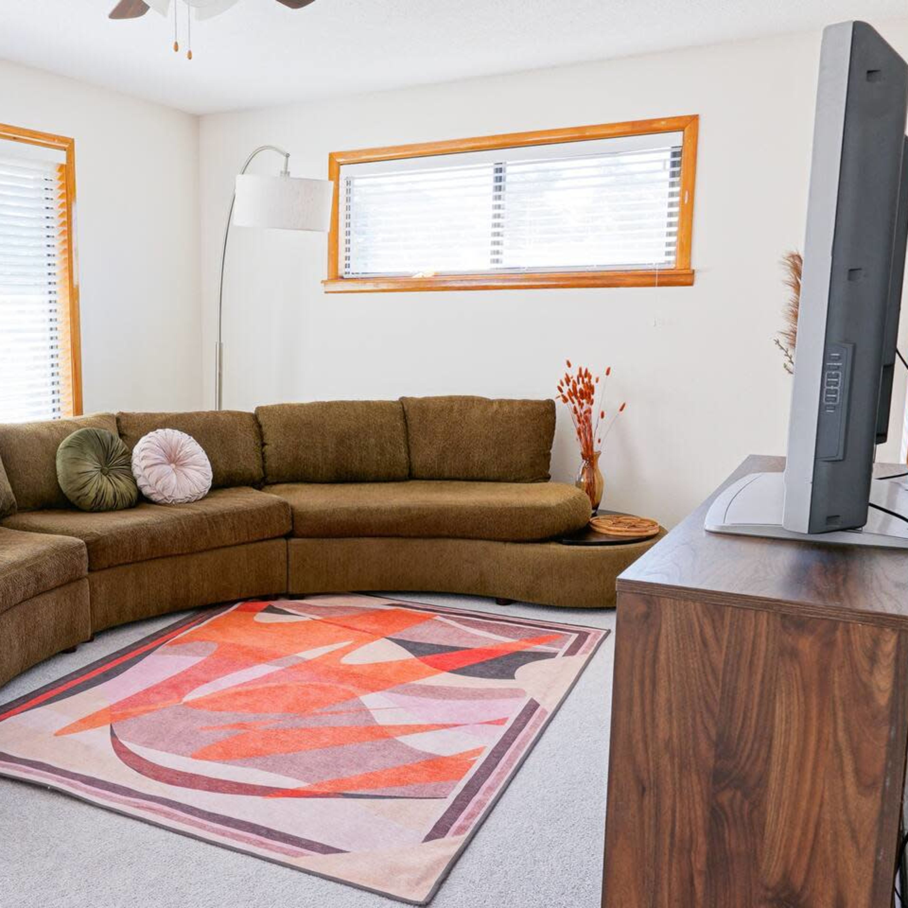 A living room with a large, curved brown sectional sofa, a light-colored area rug with geometric patterns, and a flat-screen television on a wooden stand.