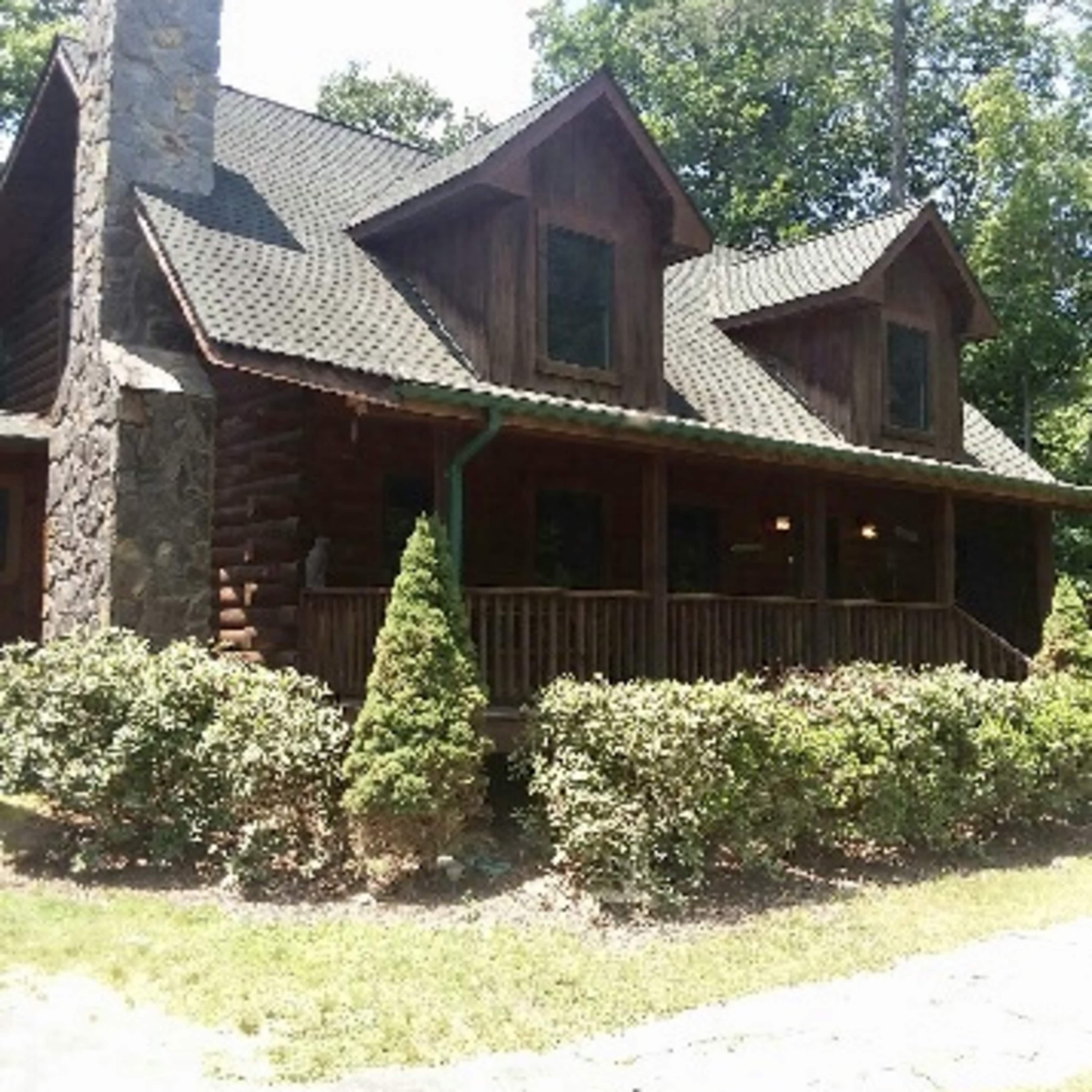 A wooden log cabin with a stone chimney and multiple gabled roofs is surrounded by greenery.