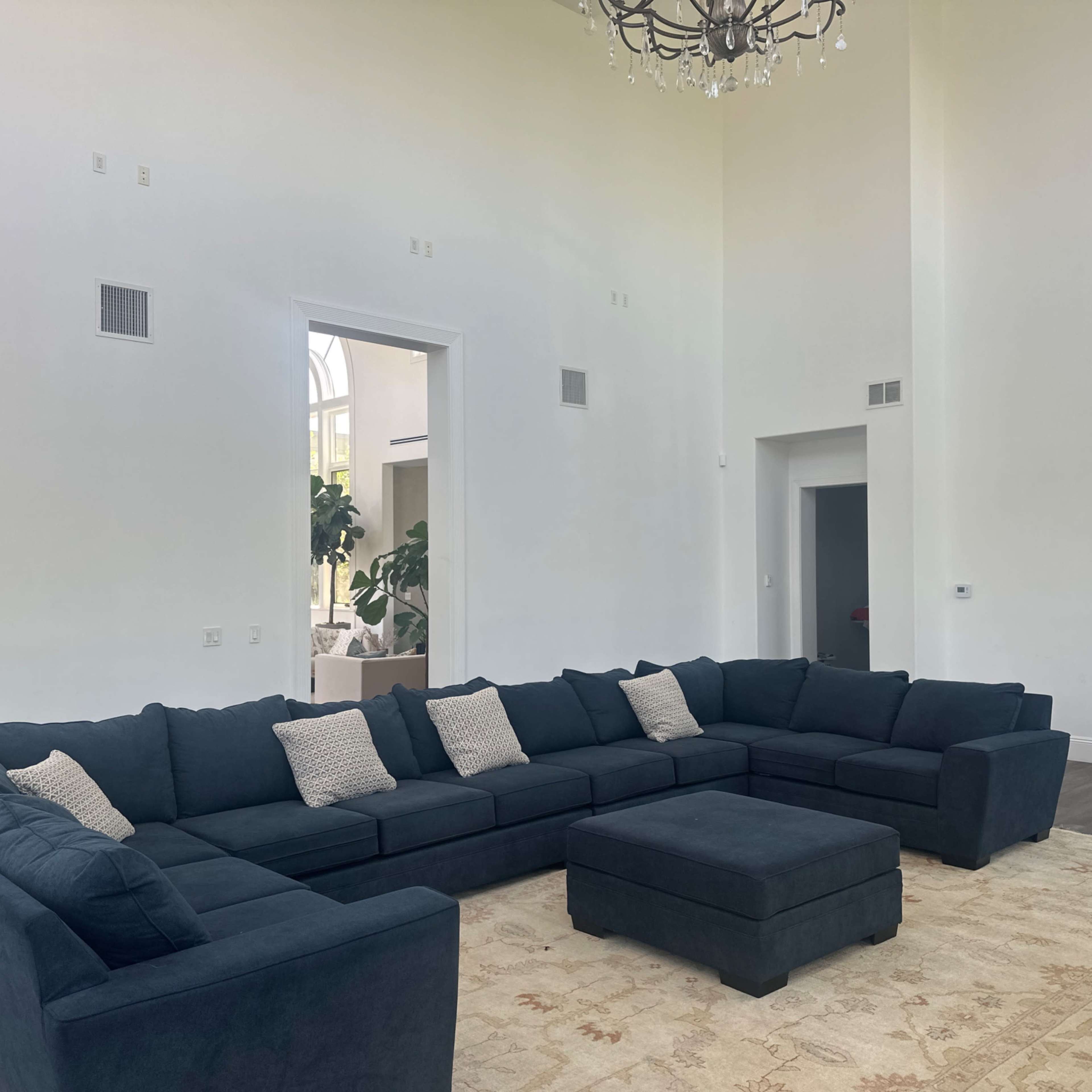 A large dark blue sectional sofa with several cushions is positioned in a spacious, brightly lit living room featuring high ceilings and light-colored walls.