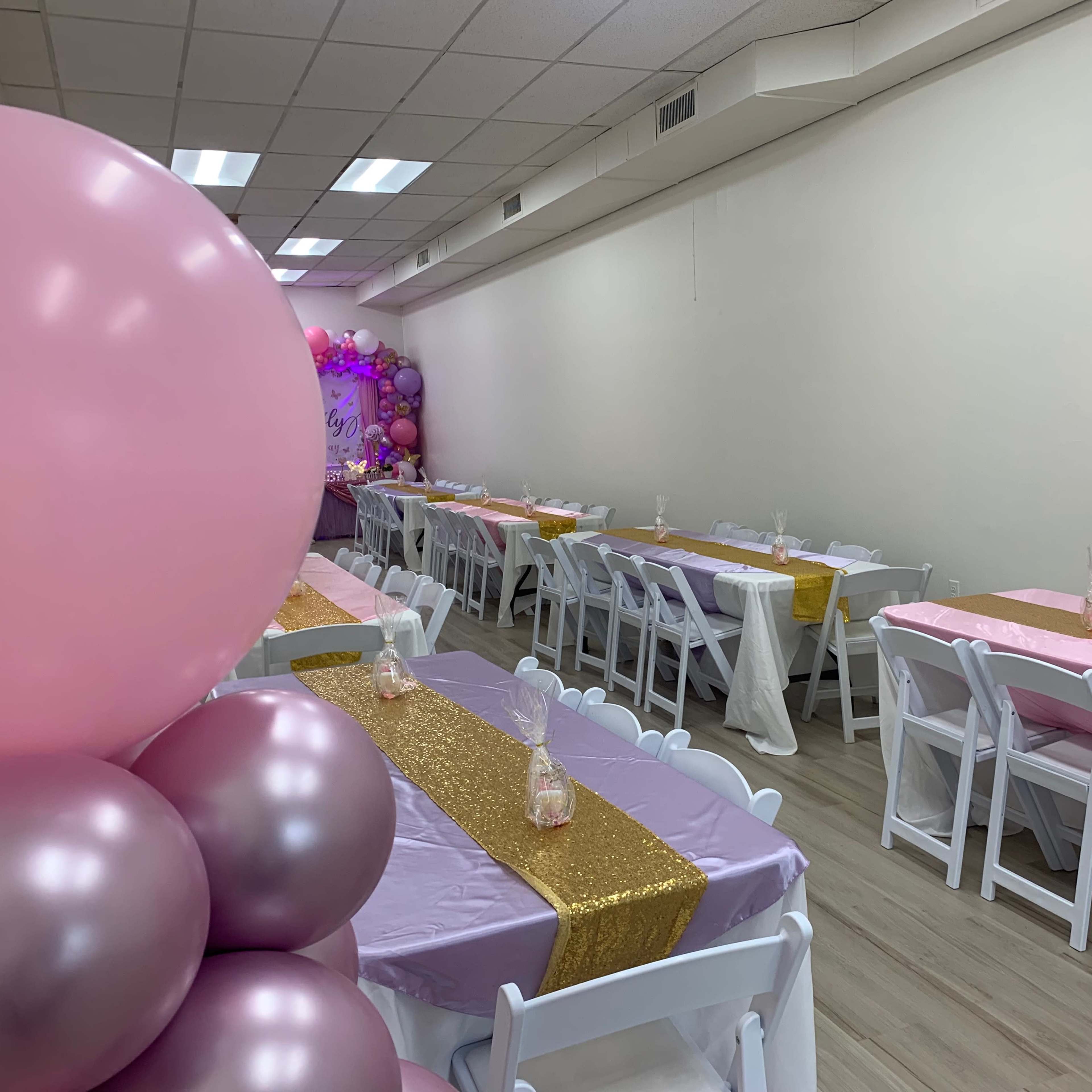 A decorated party room features tables set with pink, gold, and white tablecloths, with balloons arranged at one end.