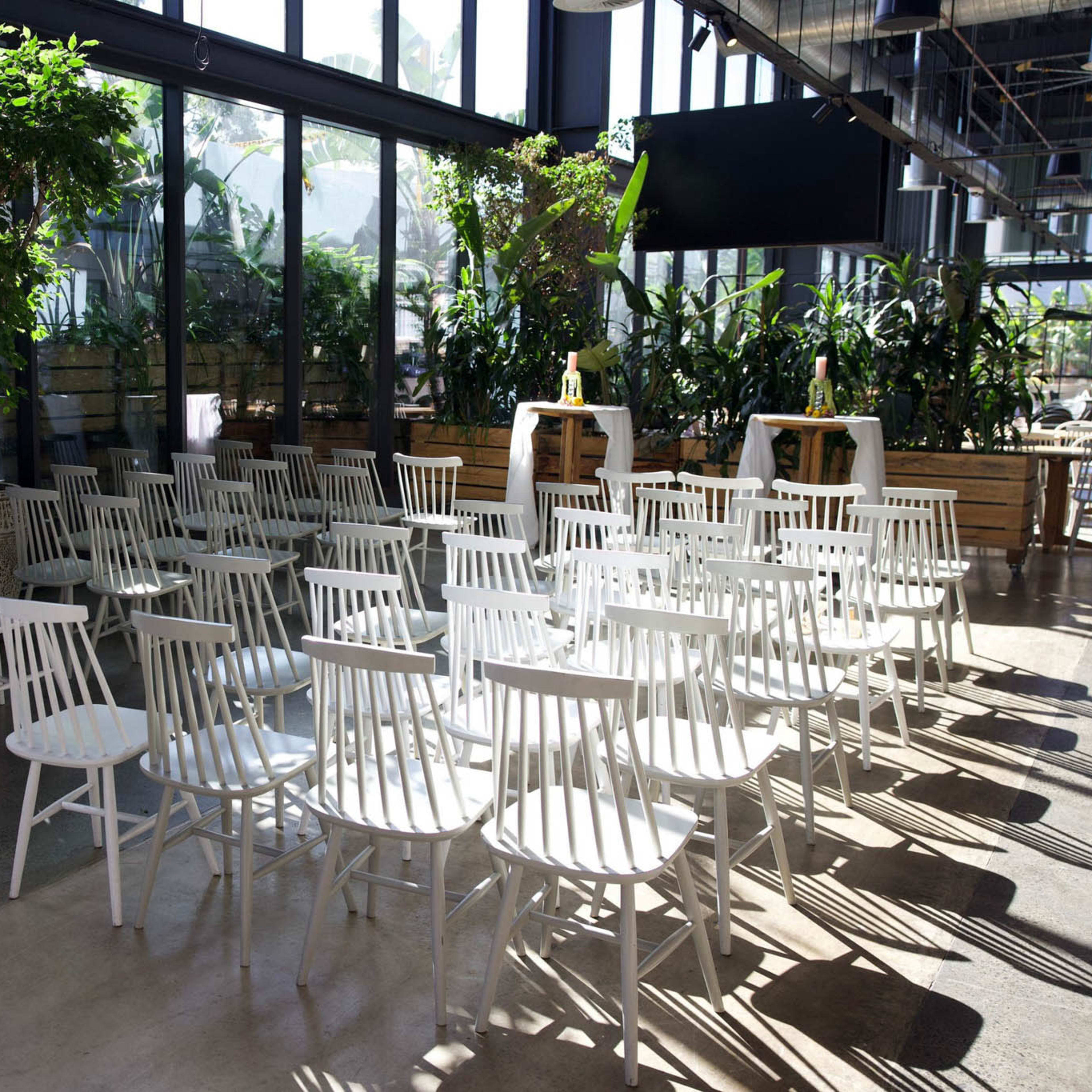 A row of white chairs is arranged in a bright, open space surrounded by large windows and greenery.