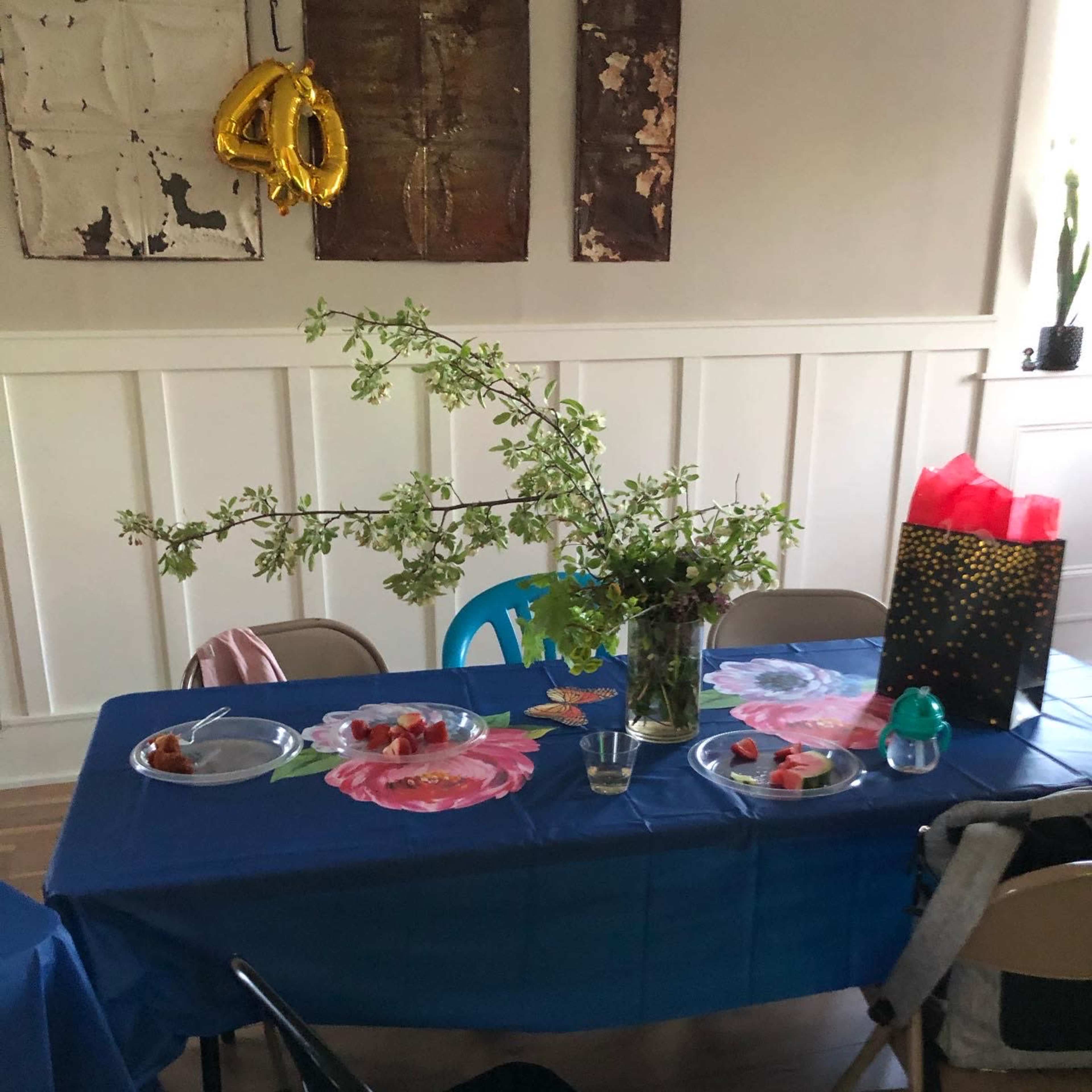 A dining table is set with blue tablecloths, decorated with floral designs, and features snacks, a large floral centerpiece, and a gift bag.