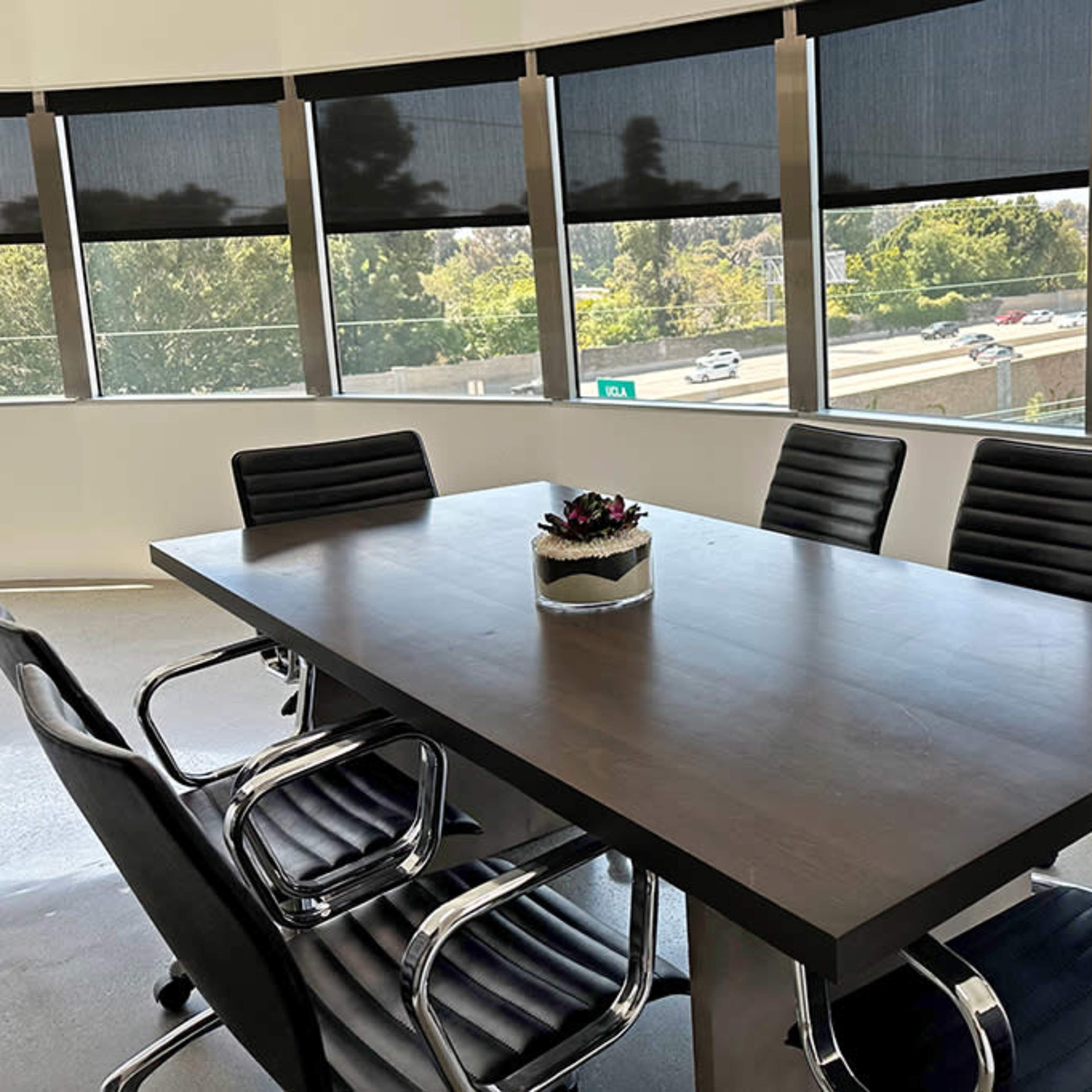 A modern conference room features a large table surrounded by black chairs, with large windows showcasing a view of a highway and greenery outside.