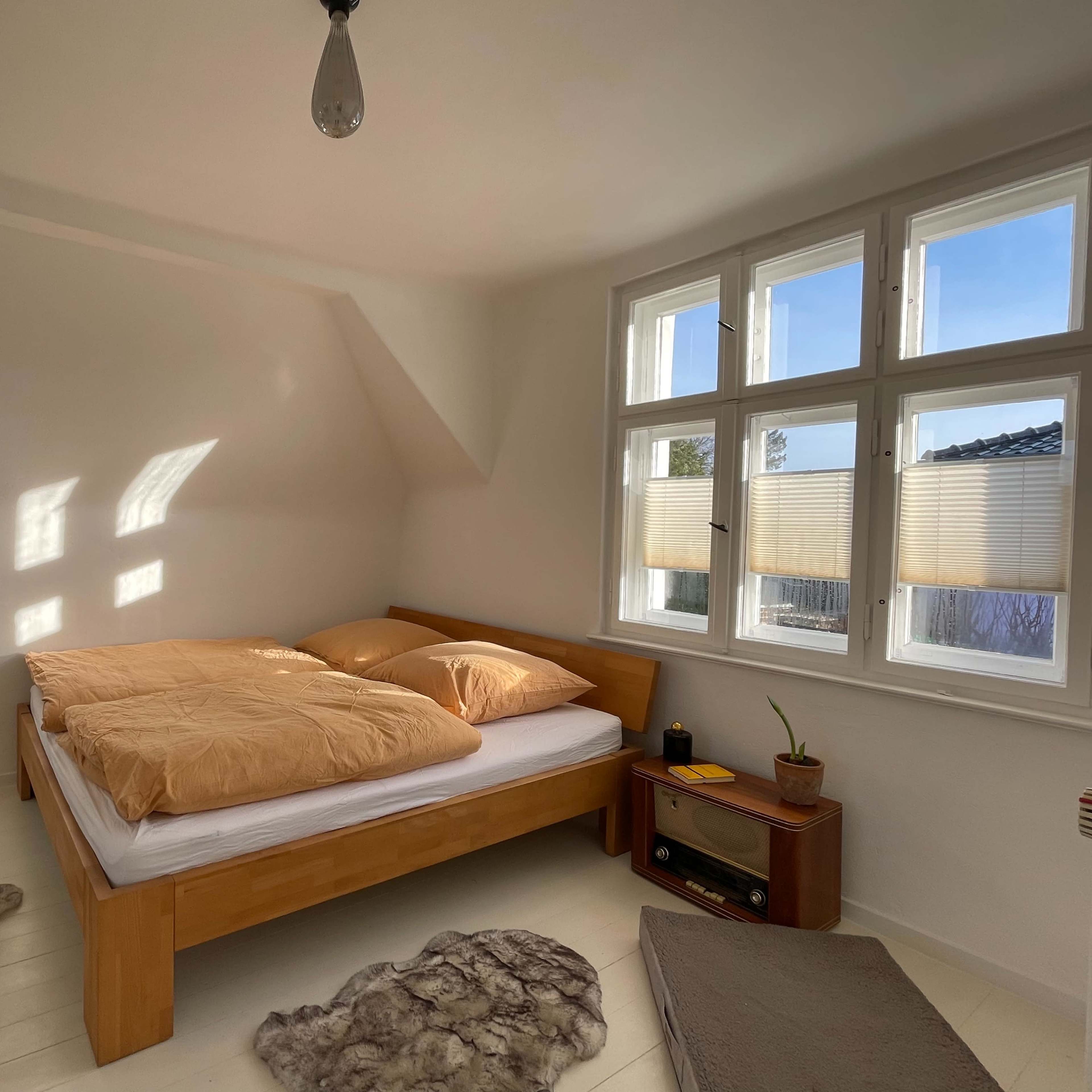 A light-filled bedroom features a wooden bed with orange bedding, a wooden side table, and two area rugs on a white floor.