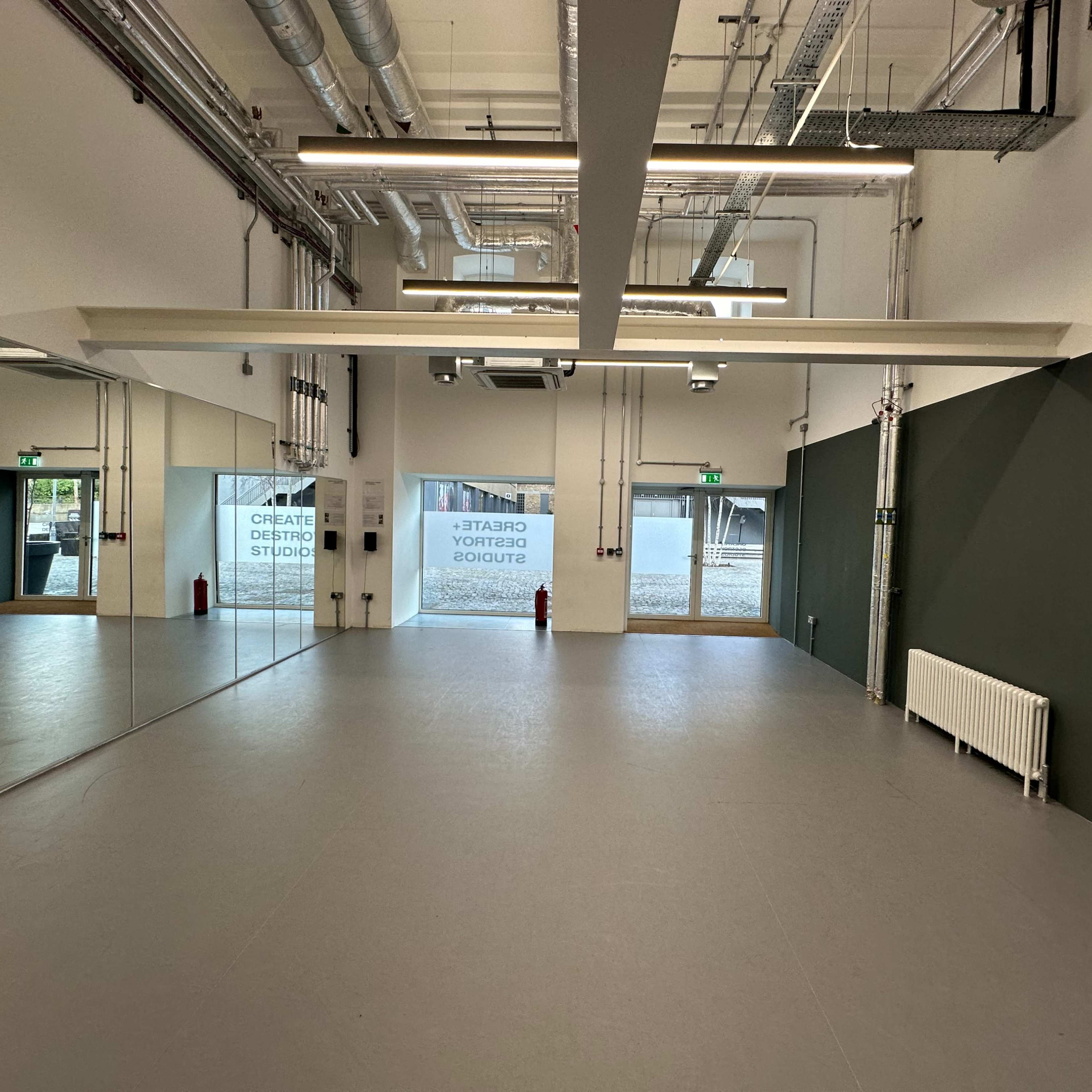 An empty studio space with mirrors on one wall, green accents, and large windows providing natural light.