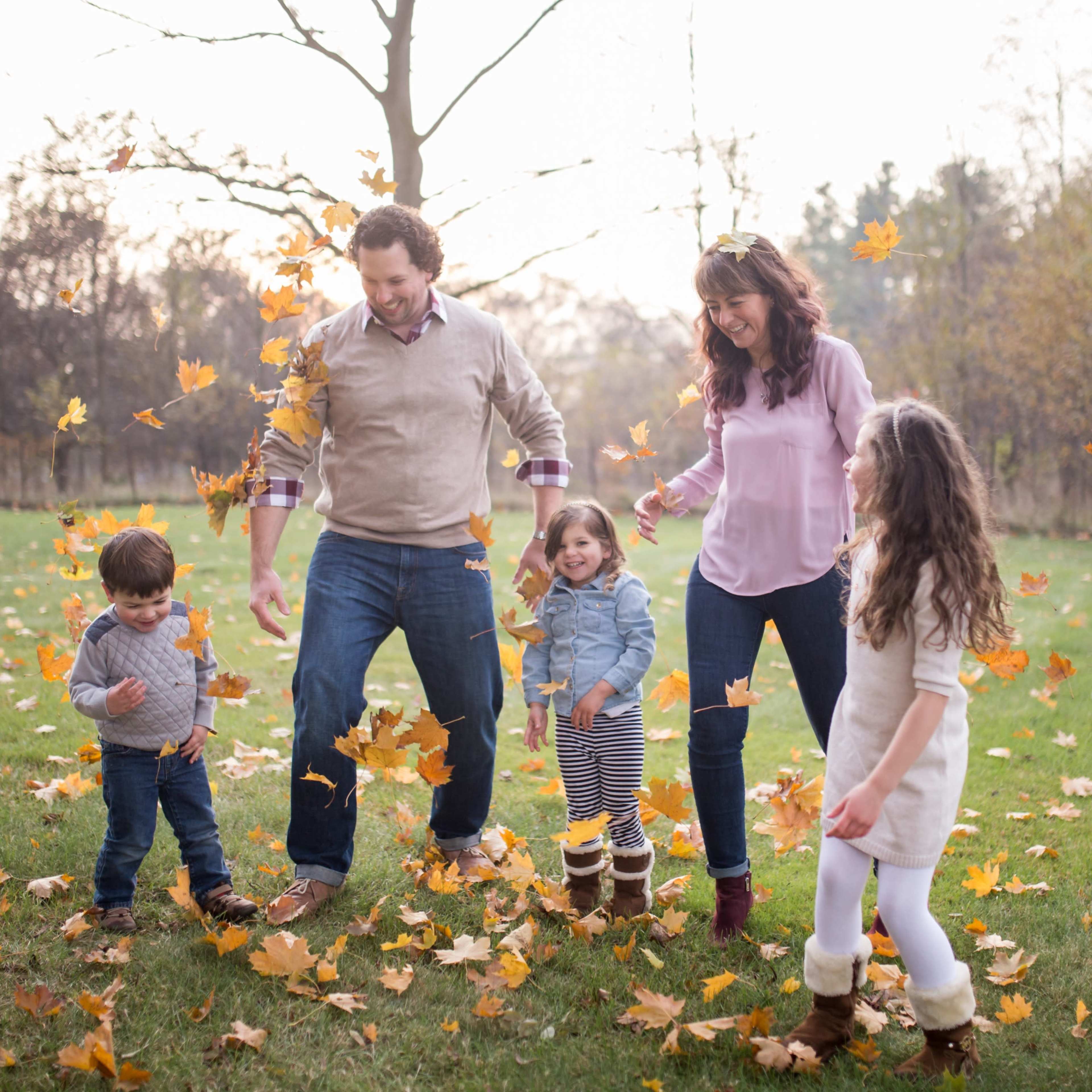 A family of five playfully tosses autumn leaves in a grassy area surrounded by trees.