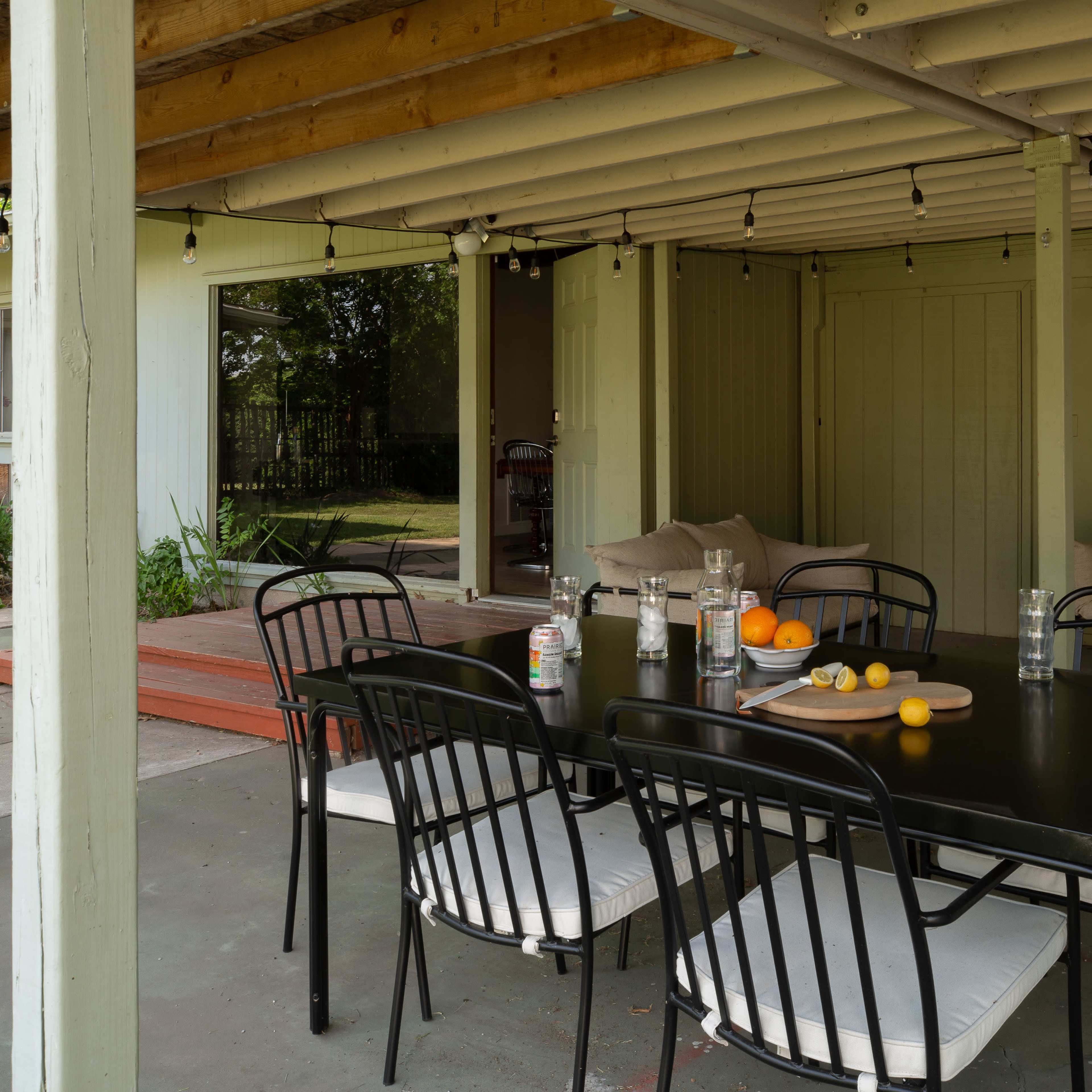 A black dining table with white cushions is set under a covered patio, surrounded by a grassy area and a wooden step leading to an outdoor space.