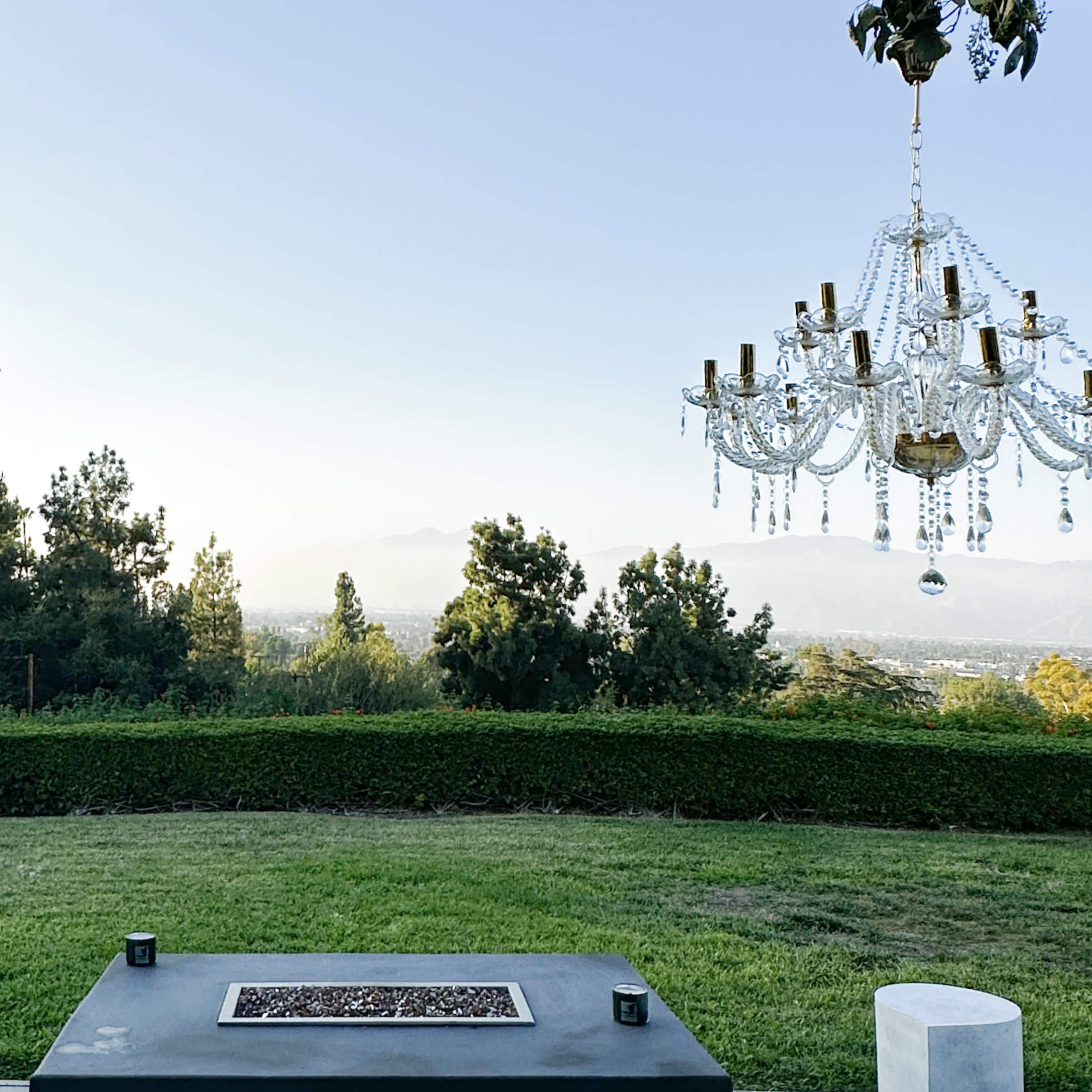 A landscaped outdoor area with a rectangular table and two ornate chandeliers hanging above, overlooking a view of distant mountains and trees.