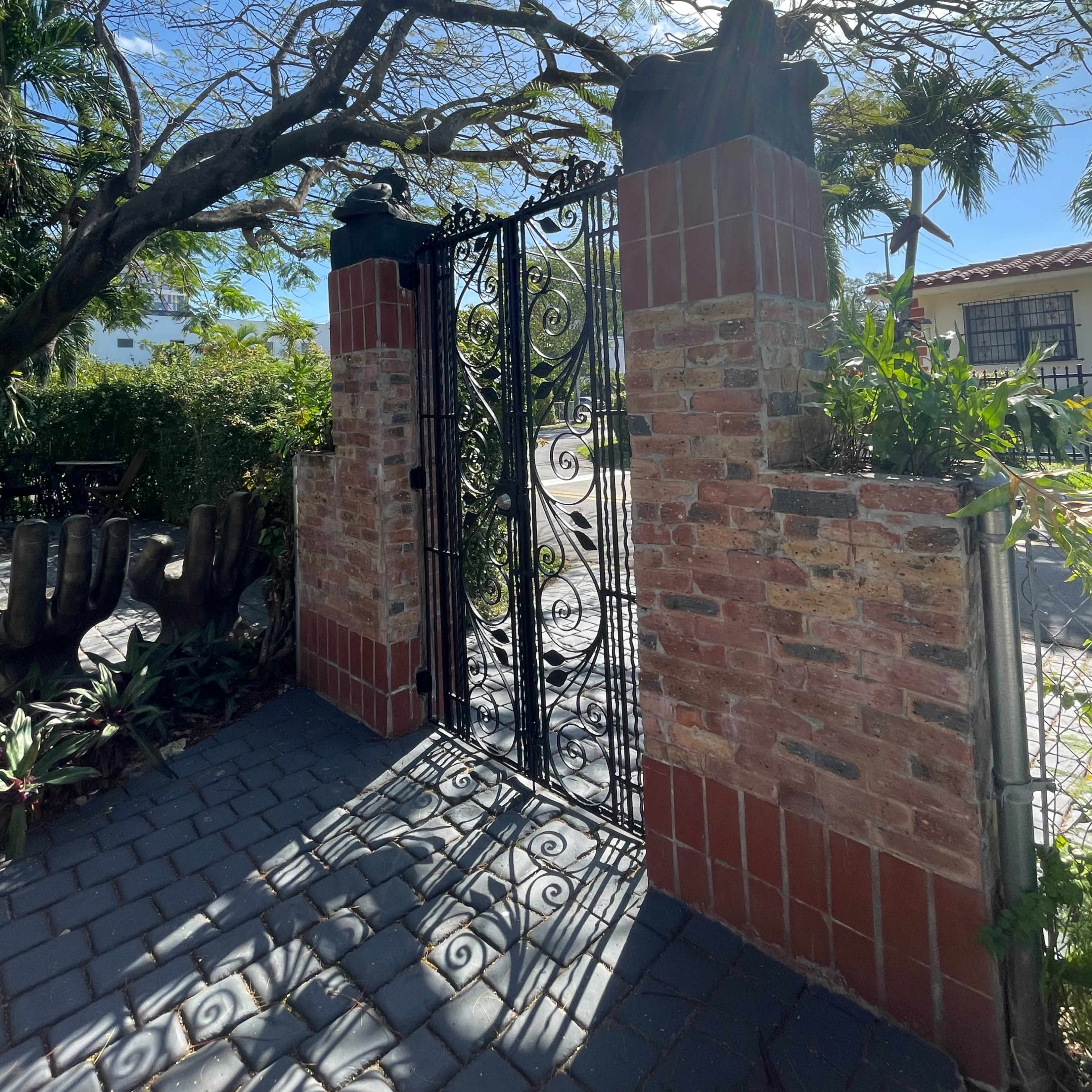 A wrought iron gate with decorative designs is flanked by brick pillars and surrounded by greenery under a bright sky.