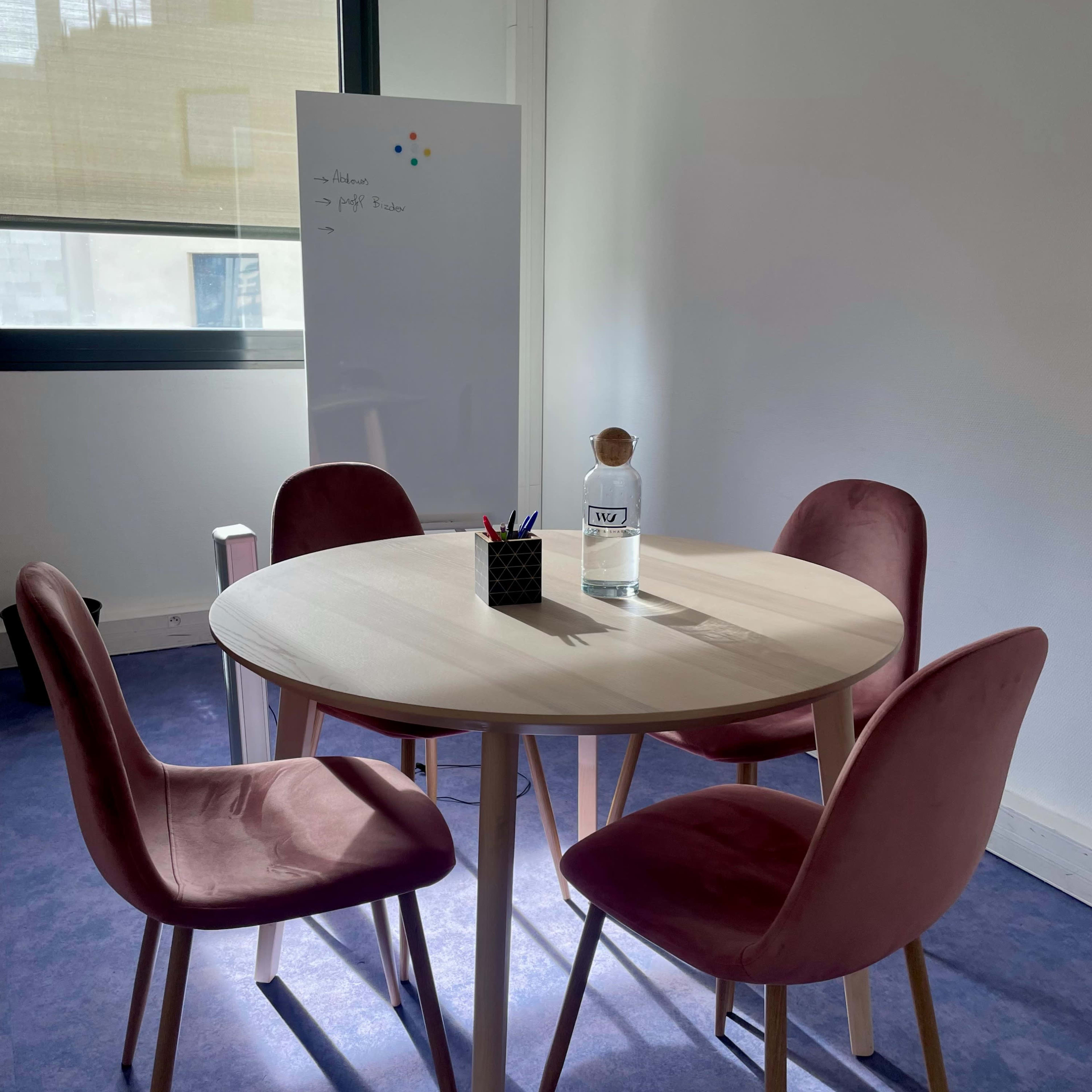 A round table with four pink chairs is set up in a bright office space next to a whiteboard.