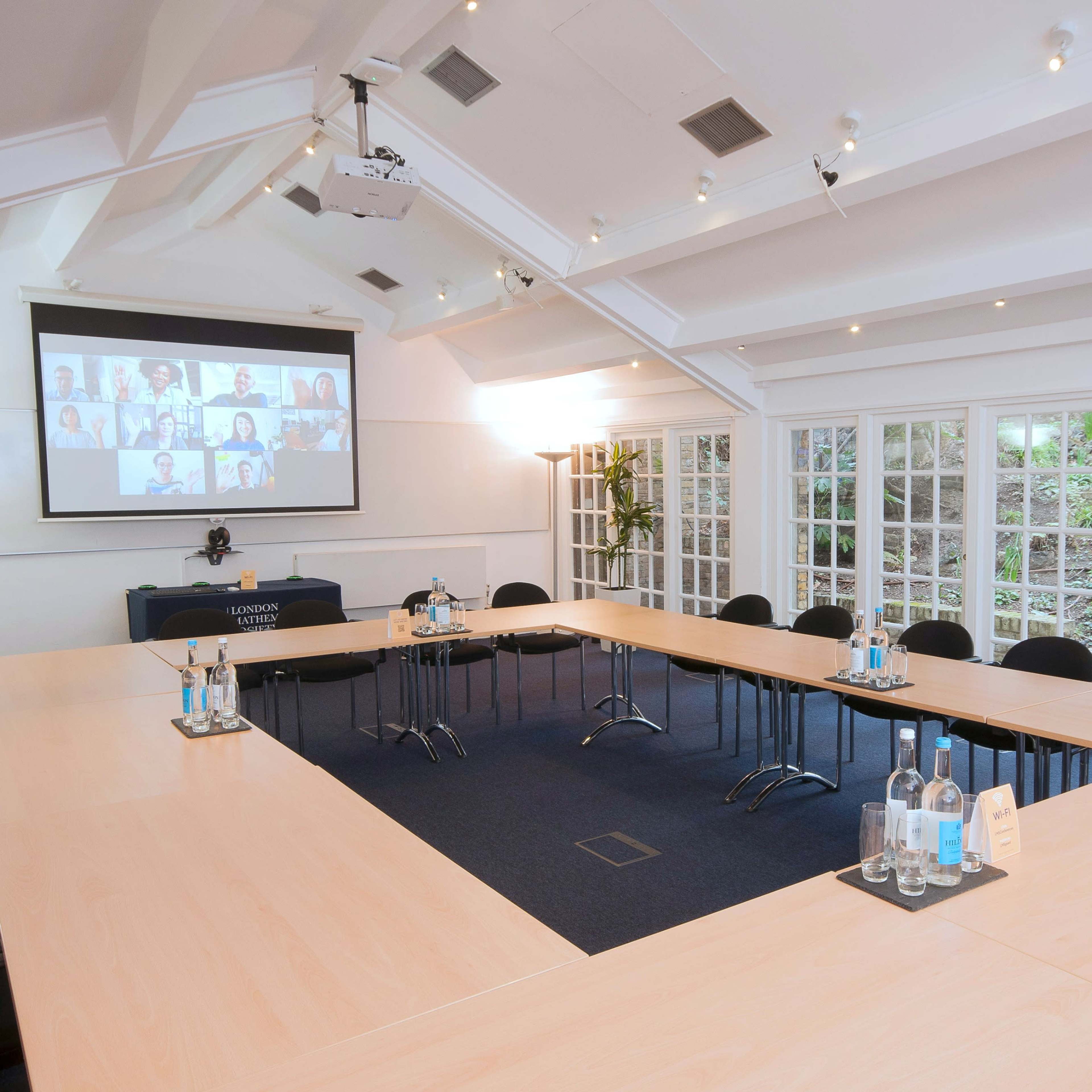 A conference room is set up with a large rectangular table, chairs arranged around it, and a screen displaying a virtual meeting in the background.