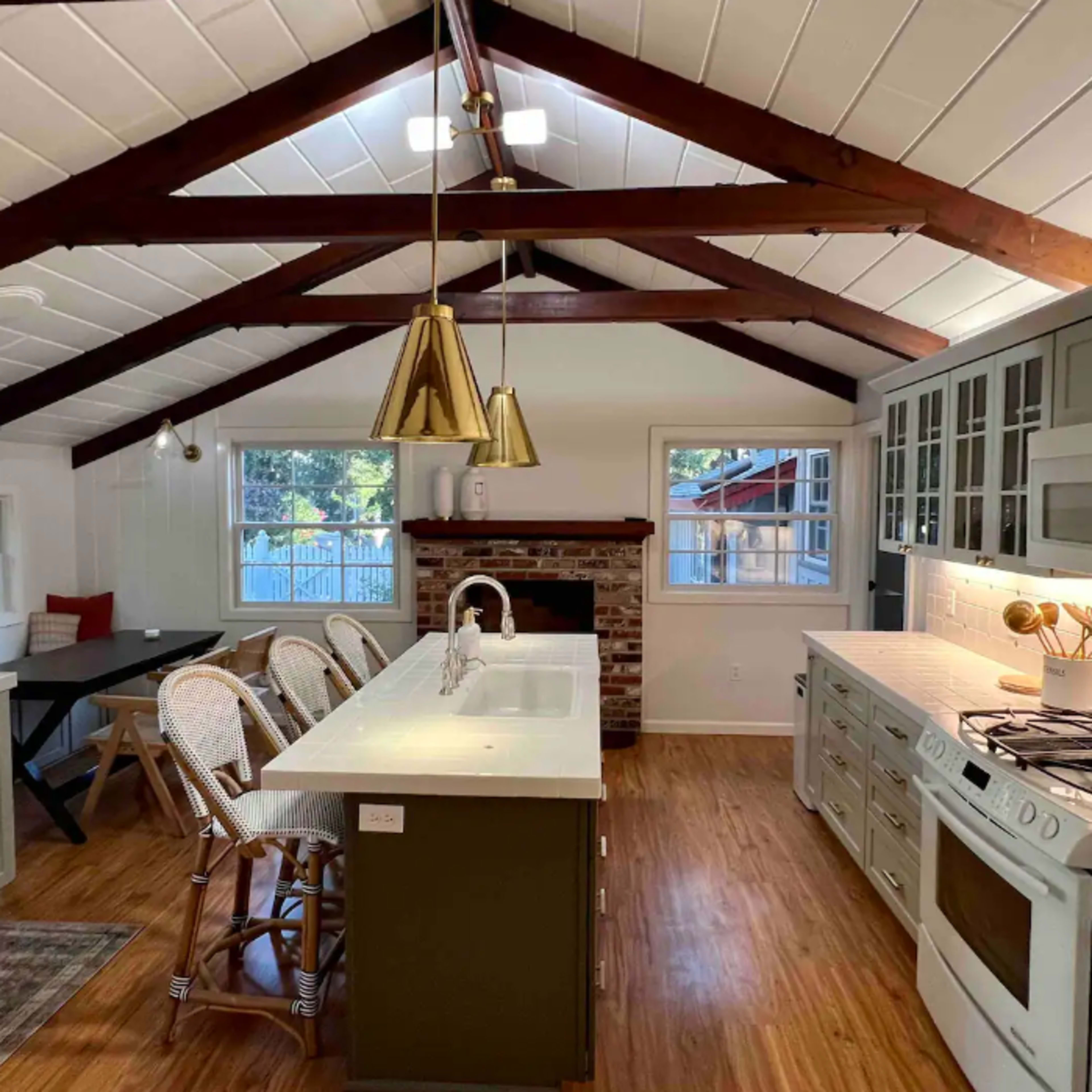 The kitchen features a high ceiling with exposed beams, a central island with seating, and modern appliances, alongside a brick fireplace.