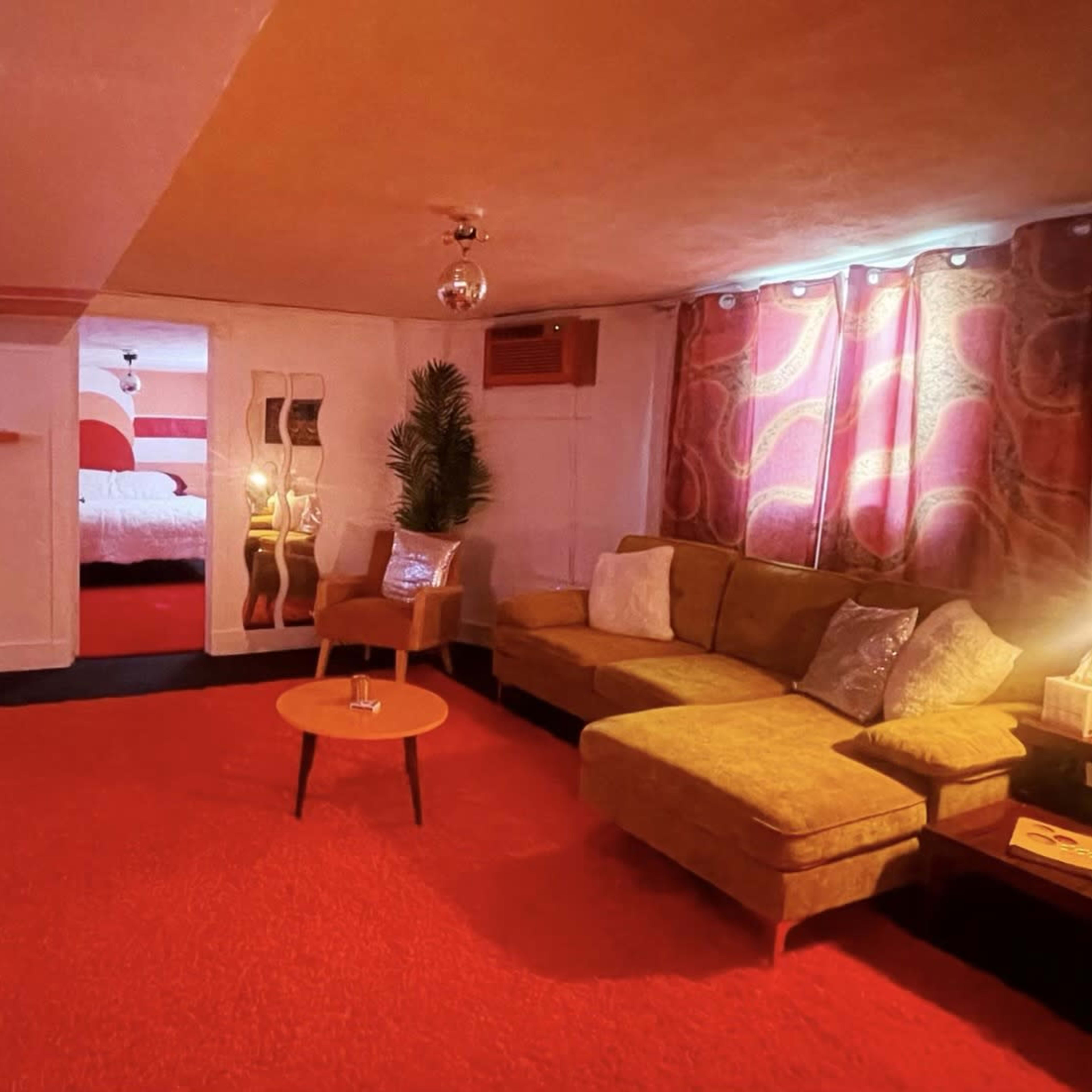 A cozy living room with a green couch, a circular coffee table, a bright orange rug, and colorful curtains, leading to a visible bedroom area.