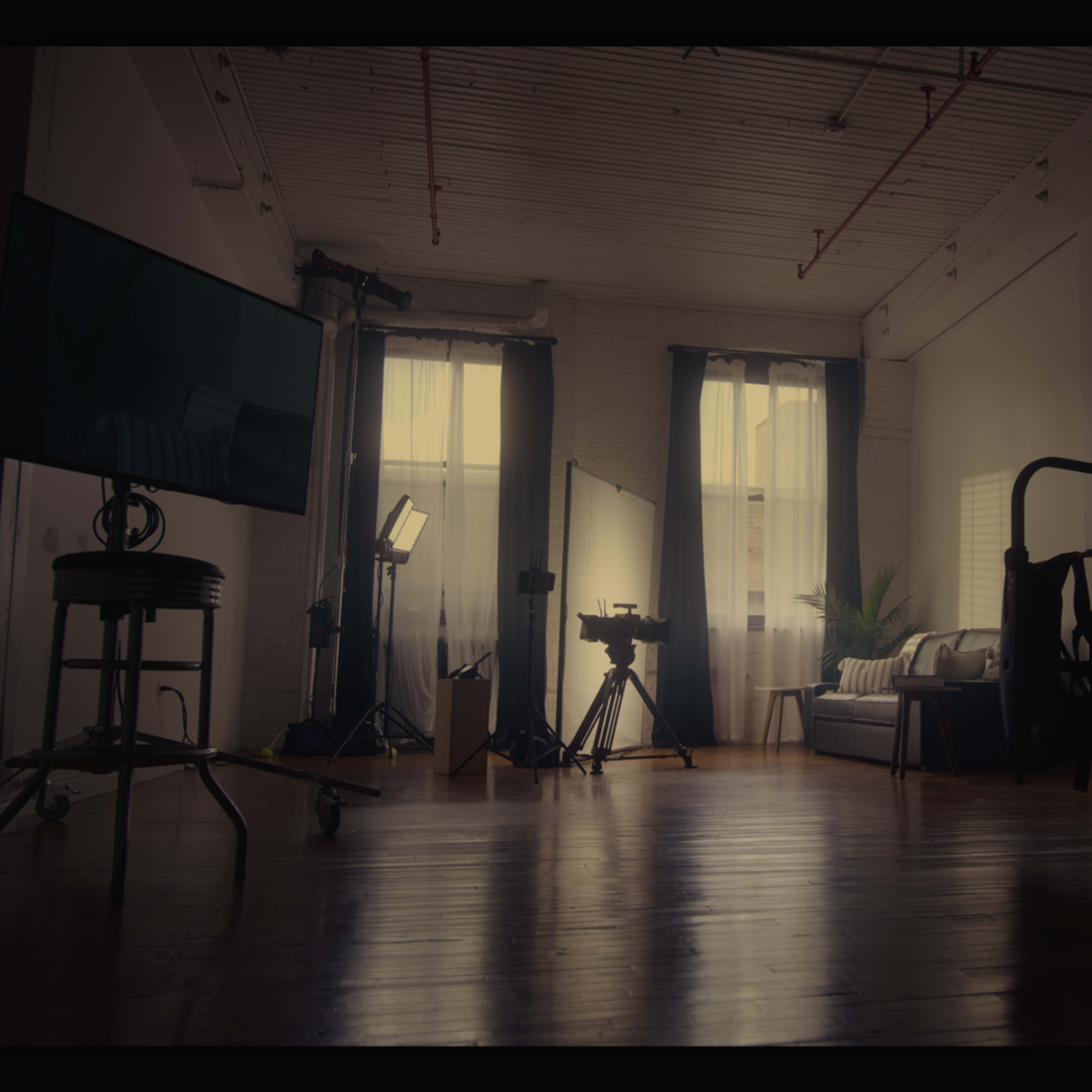 A dimly lit studio space features a sofa, lighting equipment, and camera gear arranged around a polished wooden floor.