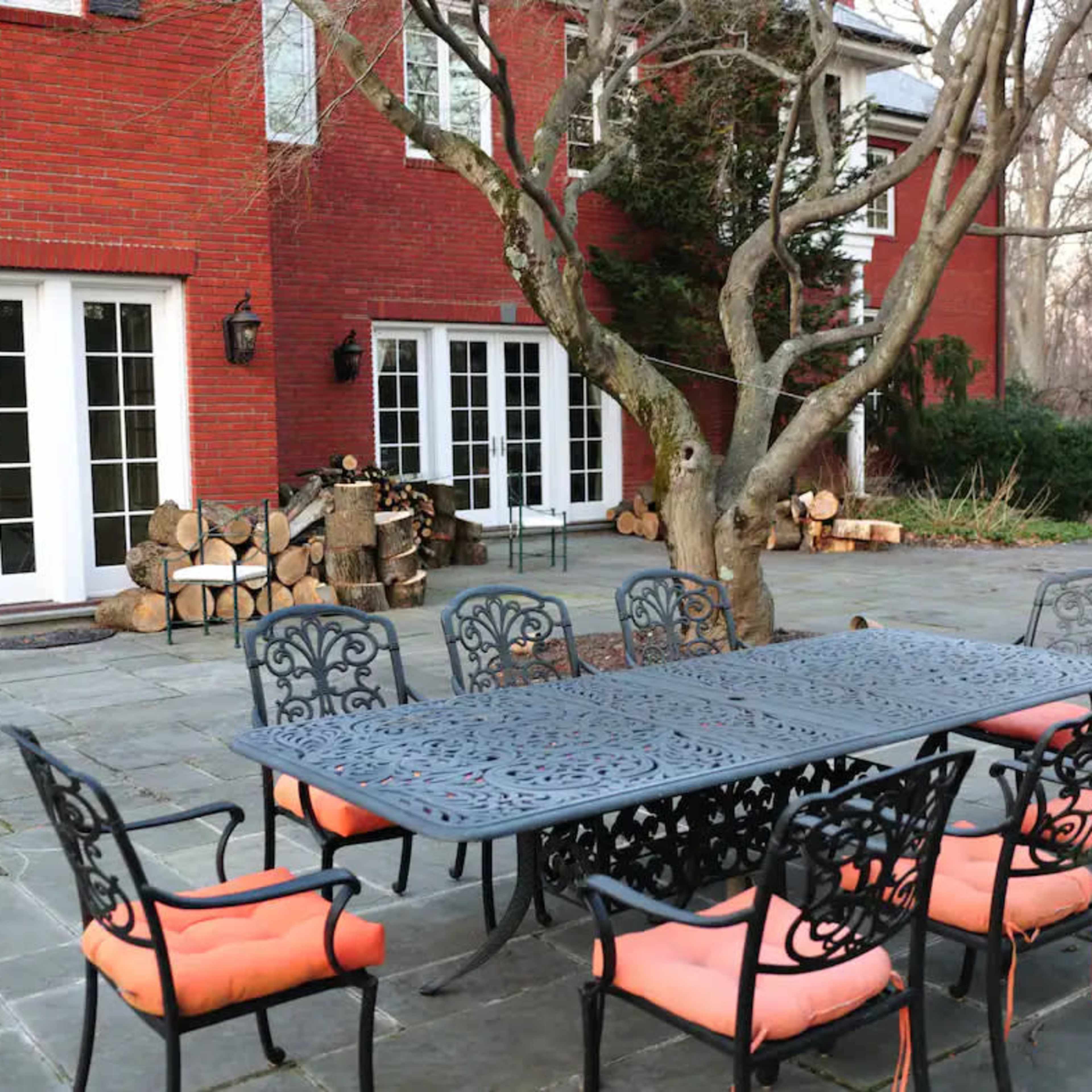 A large metal dining table with eight chairs is set on a stone patio next to a red brick house, with neatly stacked firewood in the background.