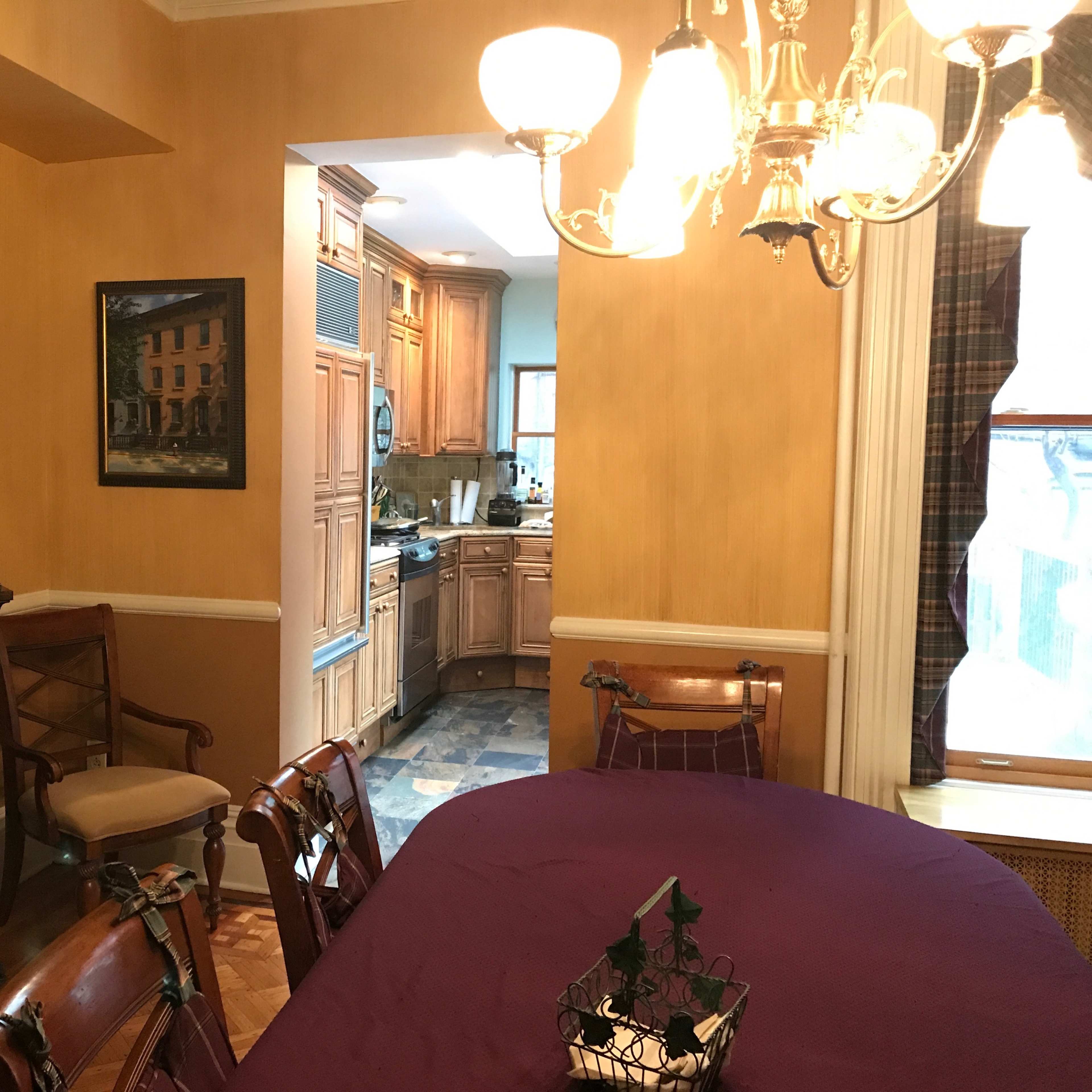 A dining room with a wooden table and chairs, adorned with a purple tablecloth, and a view into a kitchen through an open doorway.