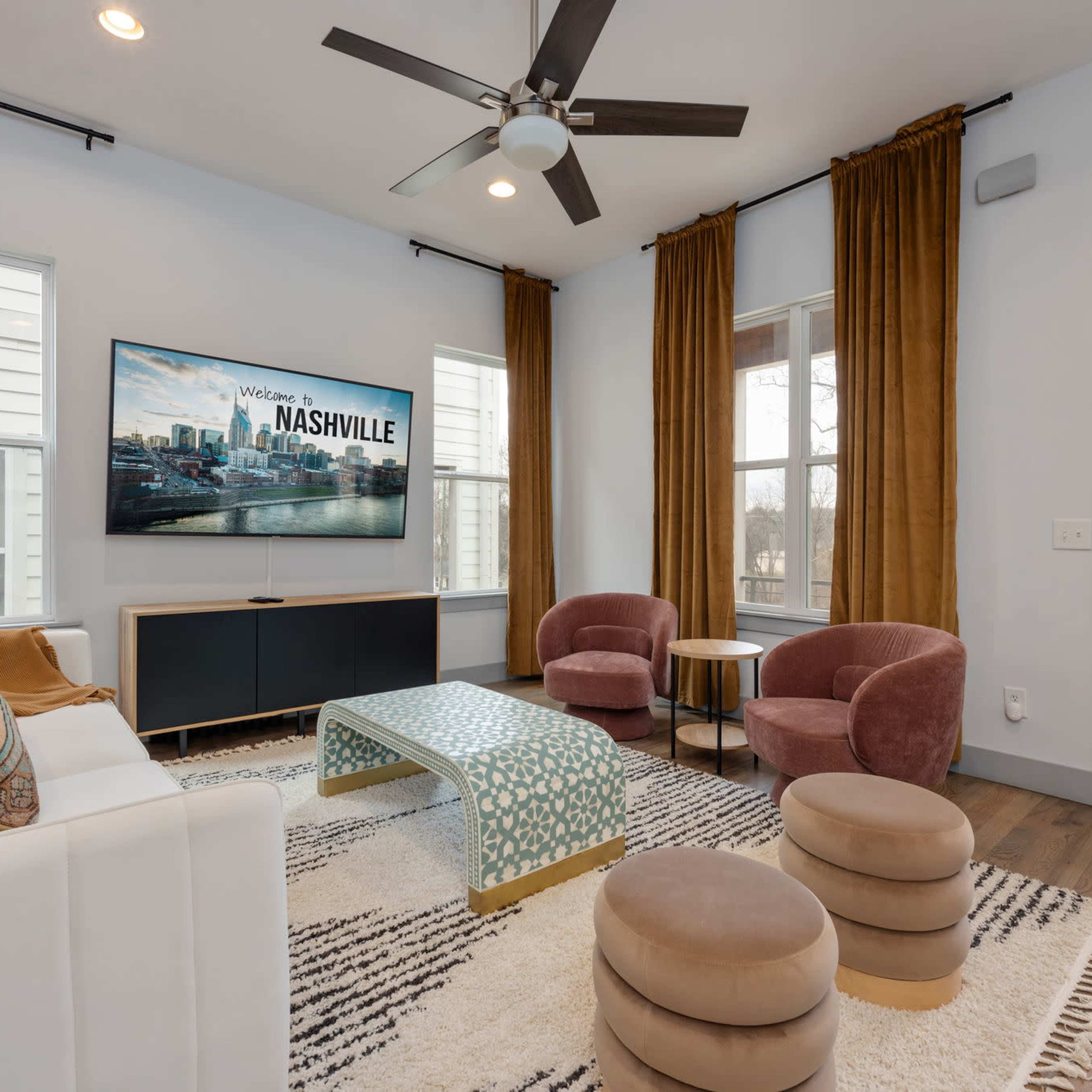 The living room features a television mounted on the wall, framed by large windows and decorated with elegant curtains, a white sofa, pink armchairs, and a patterned coffee table.