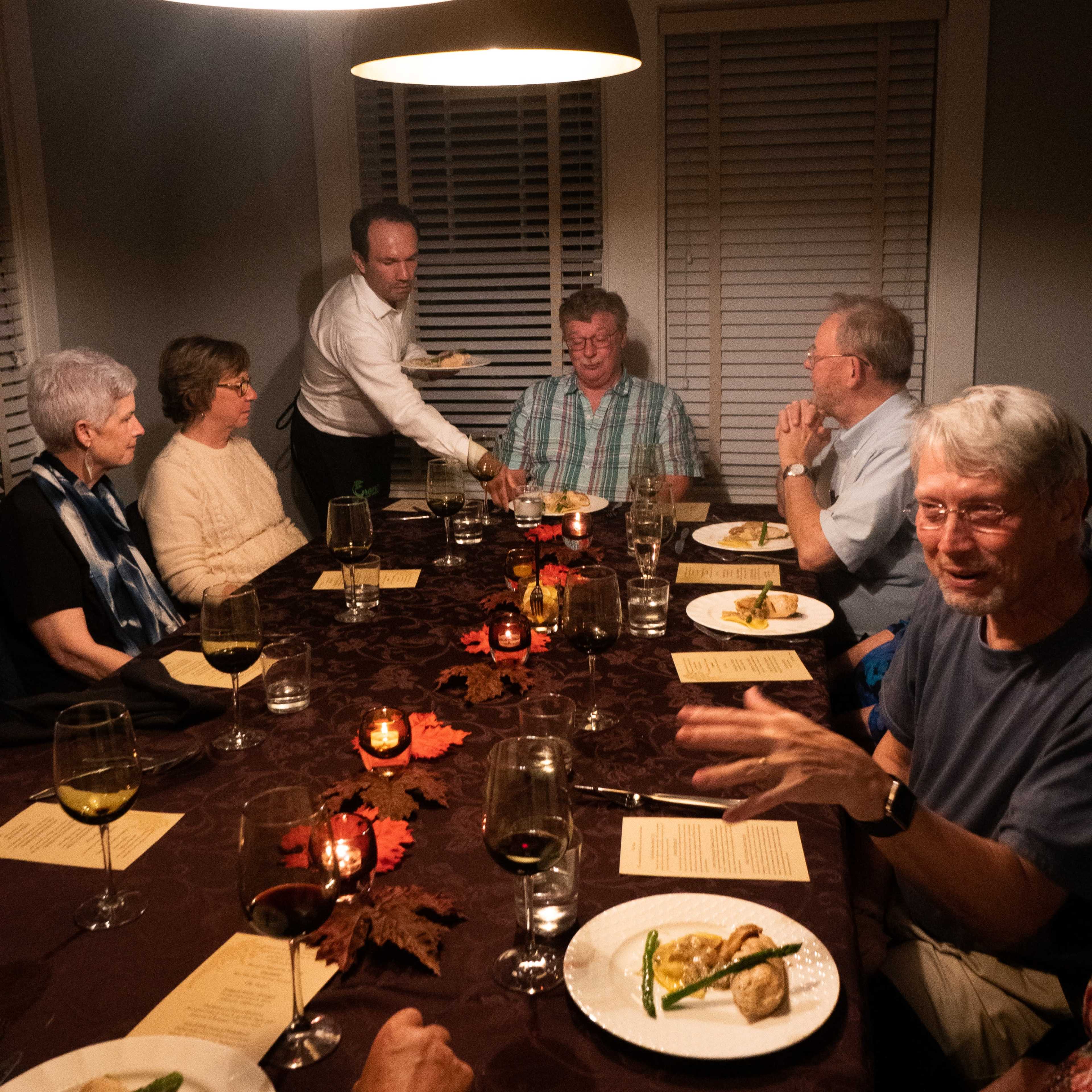 A group of eight people sit around a dining table adorned with candles and leaves as a server brings a dish to a guest.